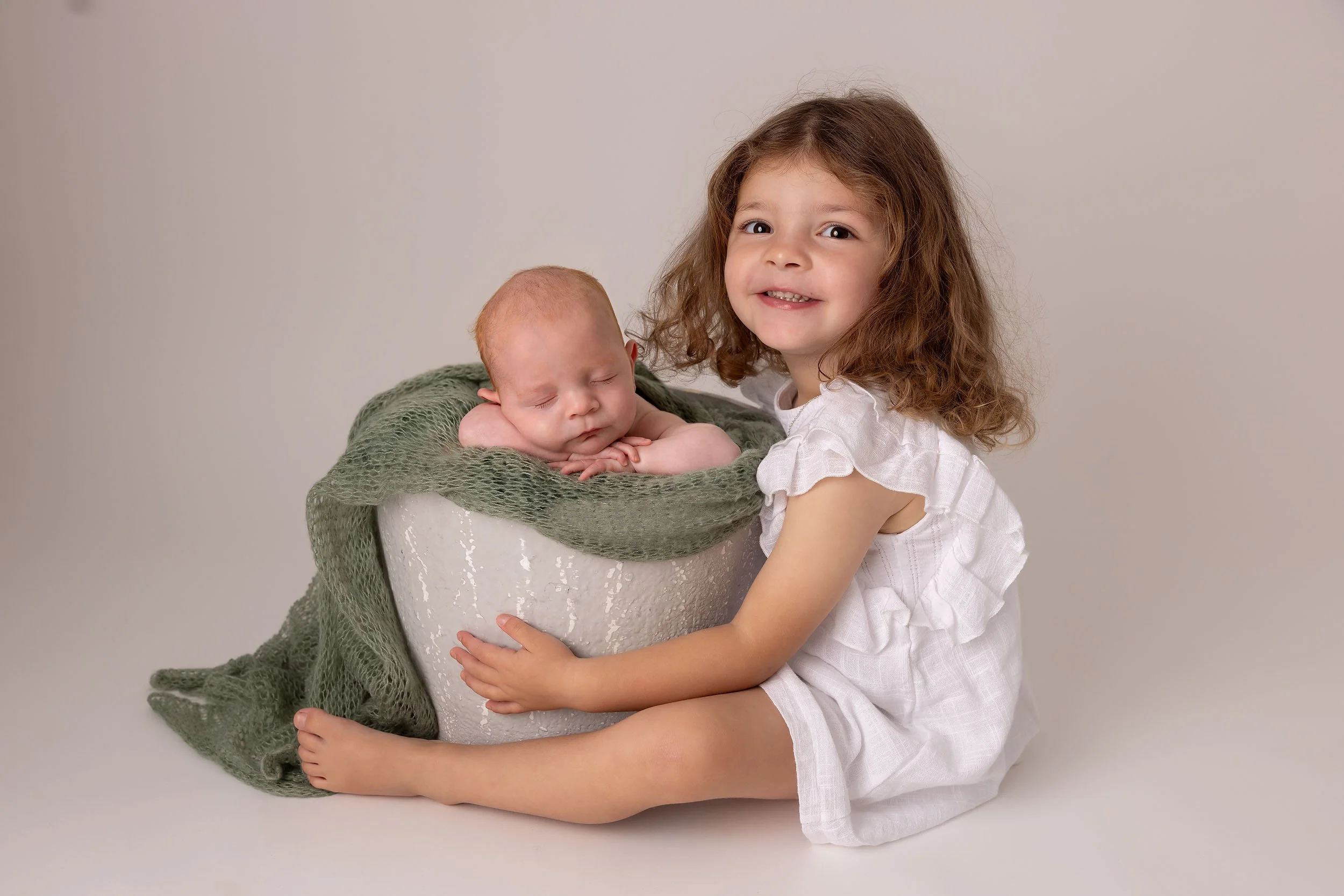 A young girl with curly brown hair and a white dress hugging a sleeping newborn baby inside a white bucket with a green blanket.