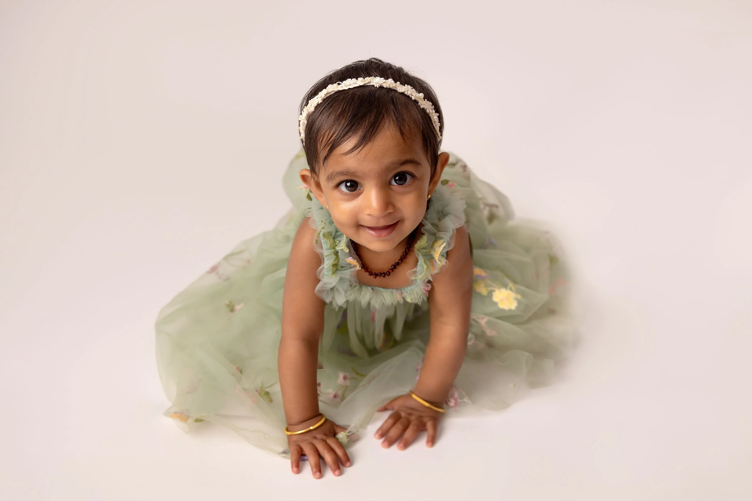 A young girl in a green floral dress kneeling on the floor, looking up and smiling at the camera.