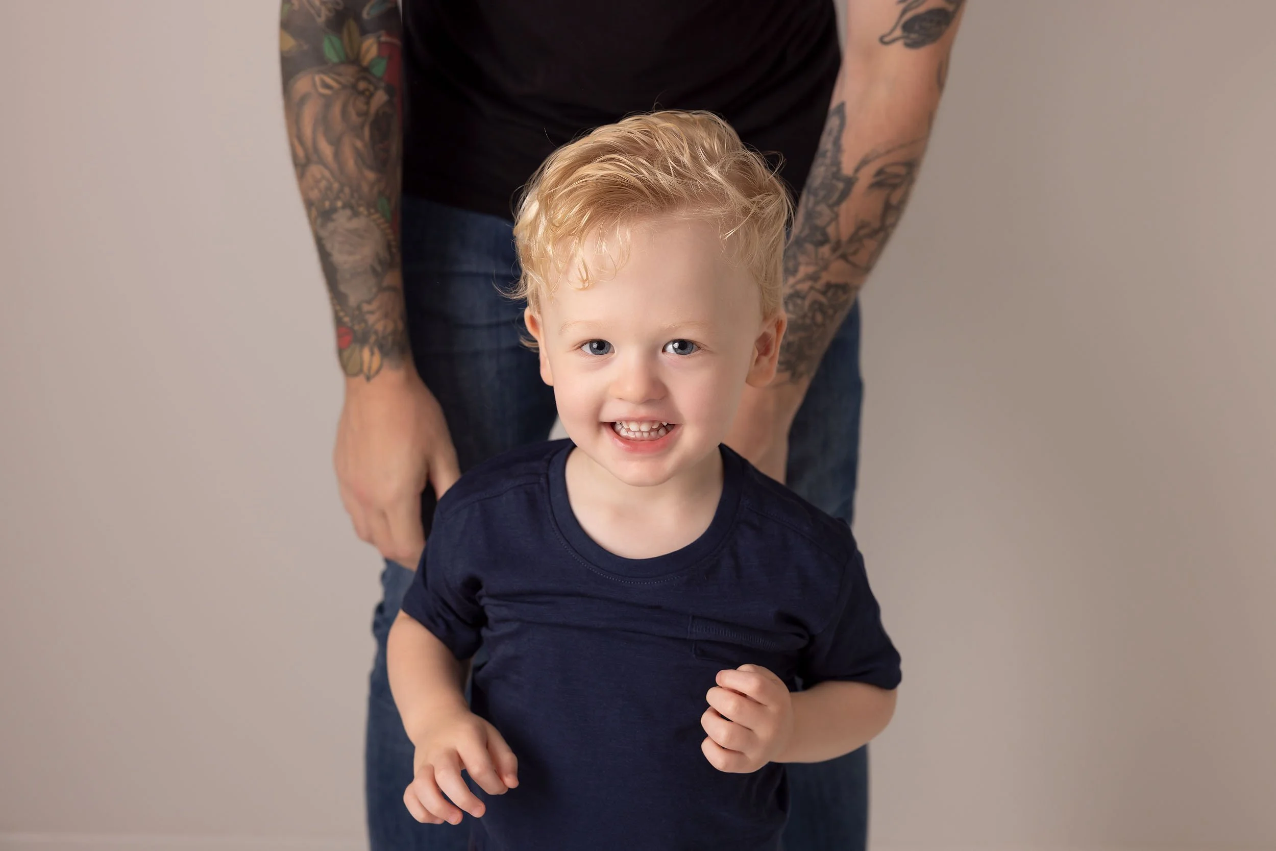 Young boy with blonde hair smiling, wearing a navy blue shirt, in front of an adult with tattooed arms.