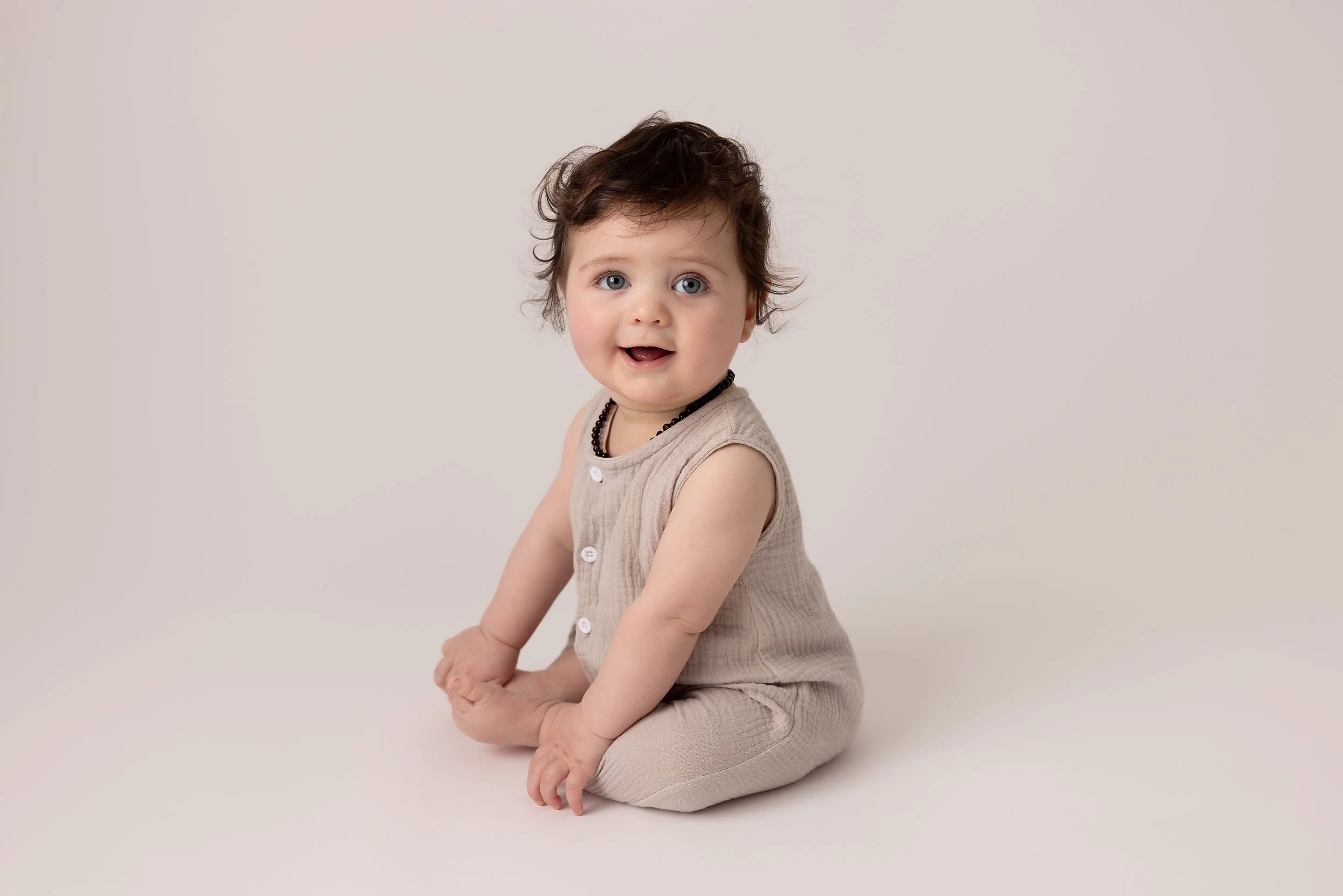 A baby with dark, curly hair sitting on a plain light background, wearing a beige sleeveless top and matching pants, with a black beaded necklace.