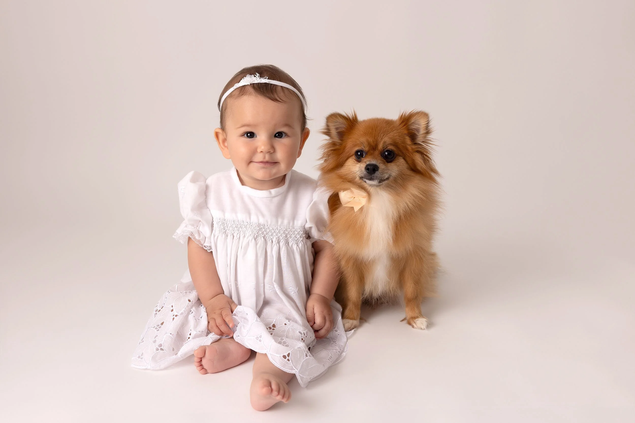 A young girl in a white dress sitting next to a small, fluffy dog with a bow around its neck against a plain background.
