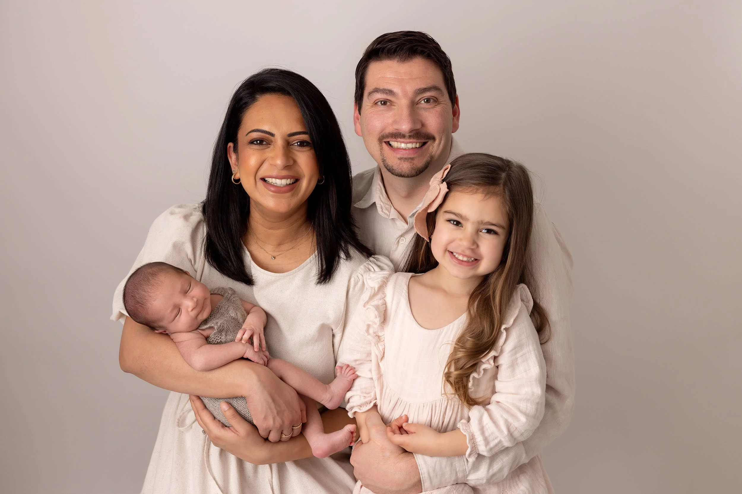Happy family of four with a newborn posing for a portrait against a plain background.