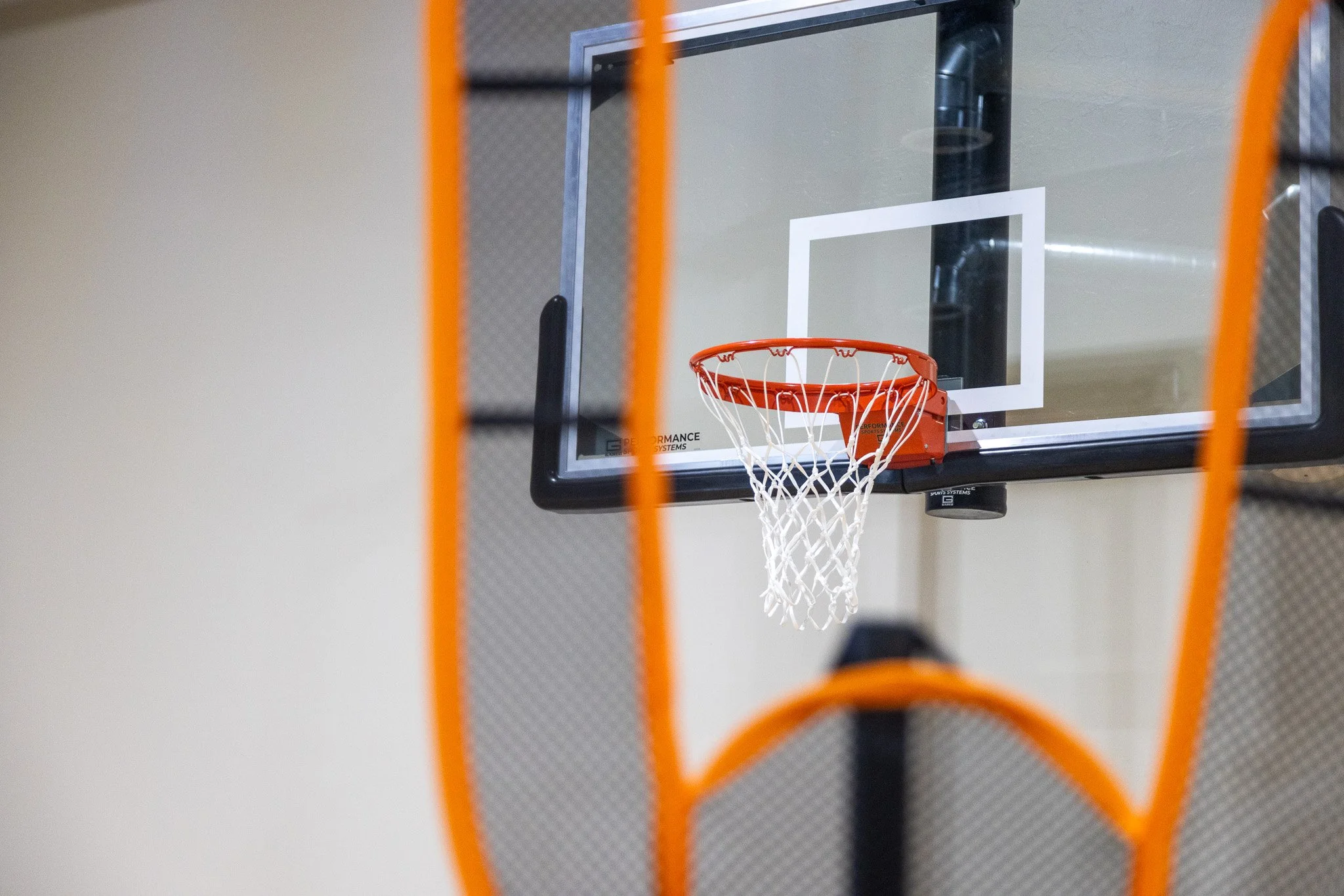 A basketball hoop with an orange rim and white net, viewed through a black and orange basketball rebounder.