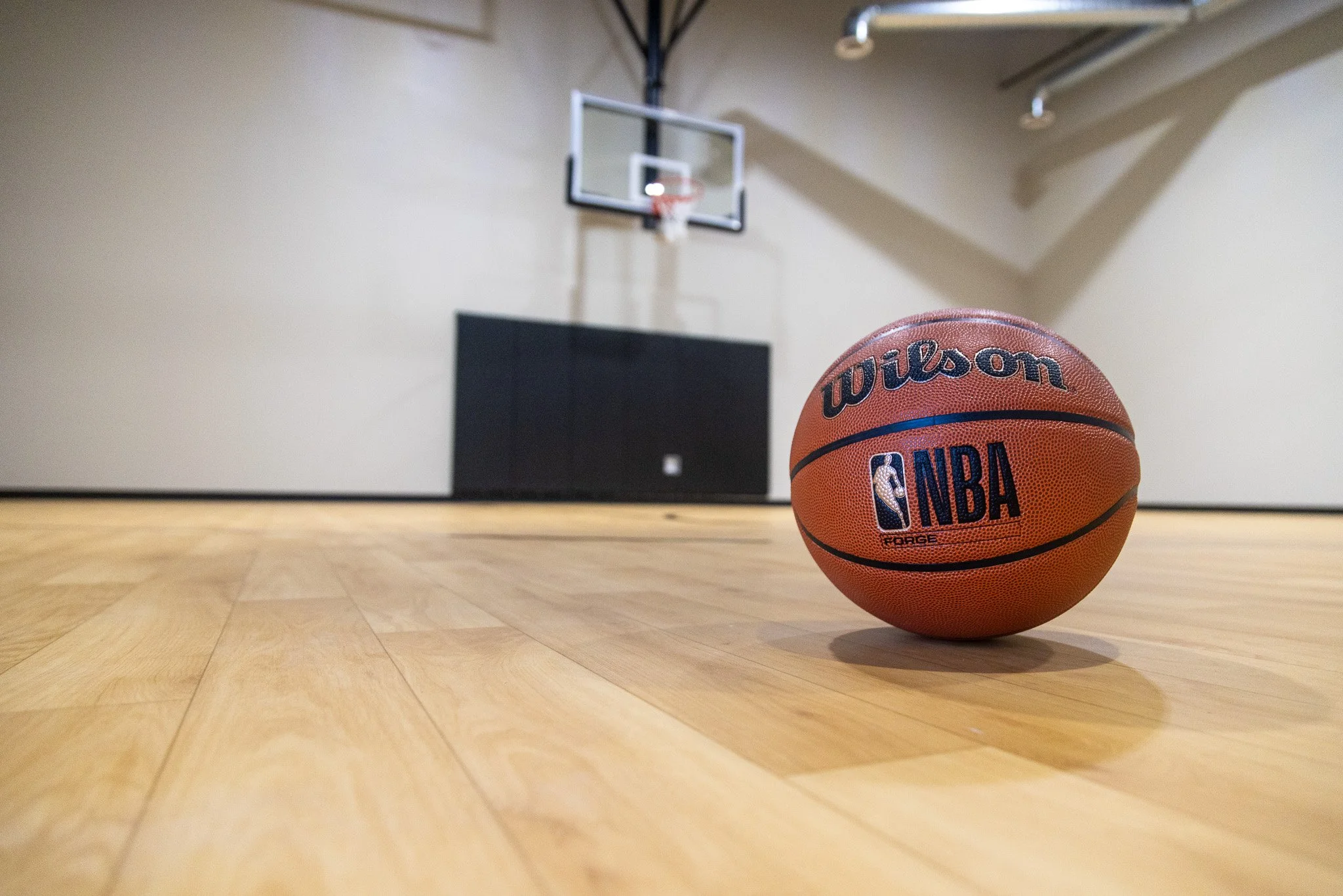 An orange Wilson NBA basketball on a wooden basketball court with a basketball hoop and backboard in the background.