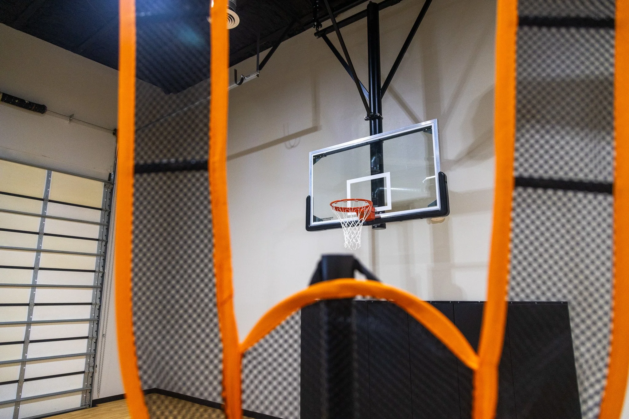 Indoor basketball court with a basketball hoop and an orange and black padded barrier in the foreground.