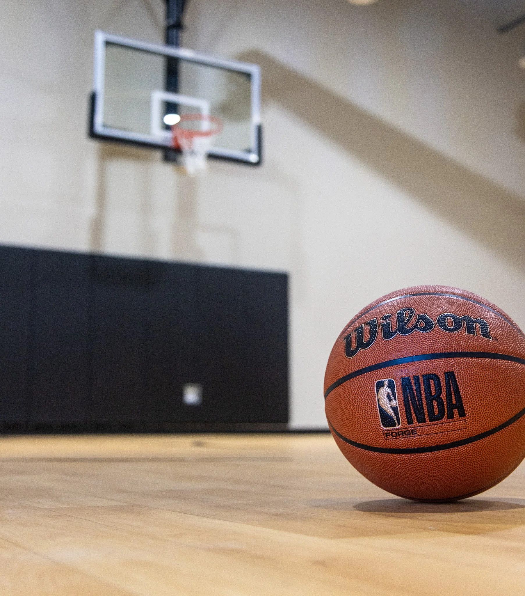 An orange Wilson NBA basketball resting on a wooden gym floor with a blurred basketball hoop in the background.