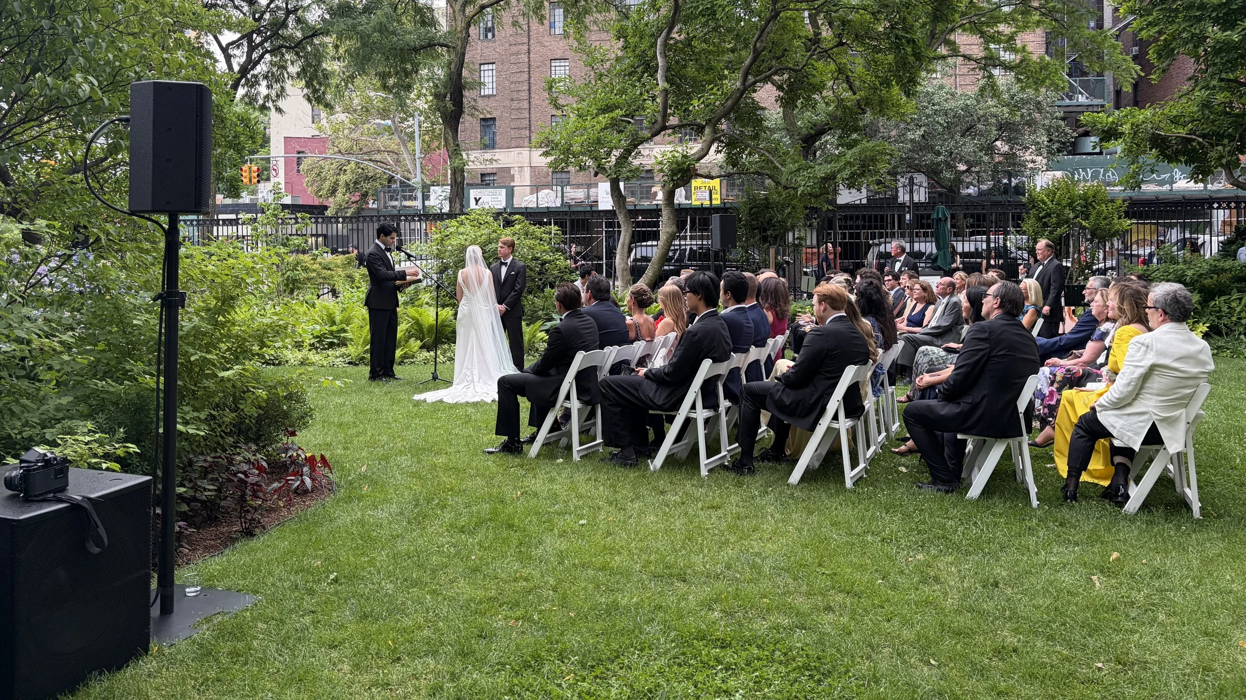 A wedding ceremony taking place outdoors in a lush green park, with a bride and groom standing under a tree, surrounded by seated guests and a speaker system.