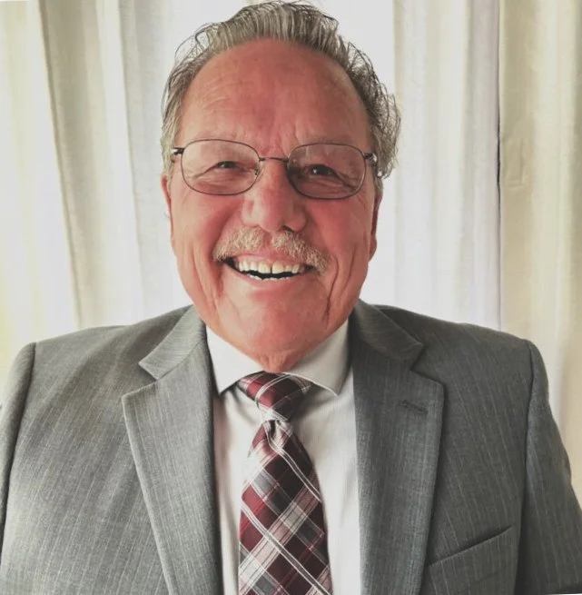 A smiling elderly man with gray hair, glasses, and a mustache, wearing a gray suit, white shirt, and plaid tie, standing in front of cream-colored curtains.