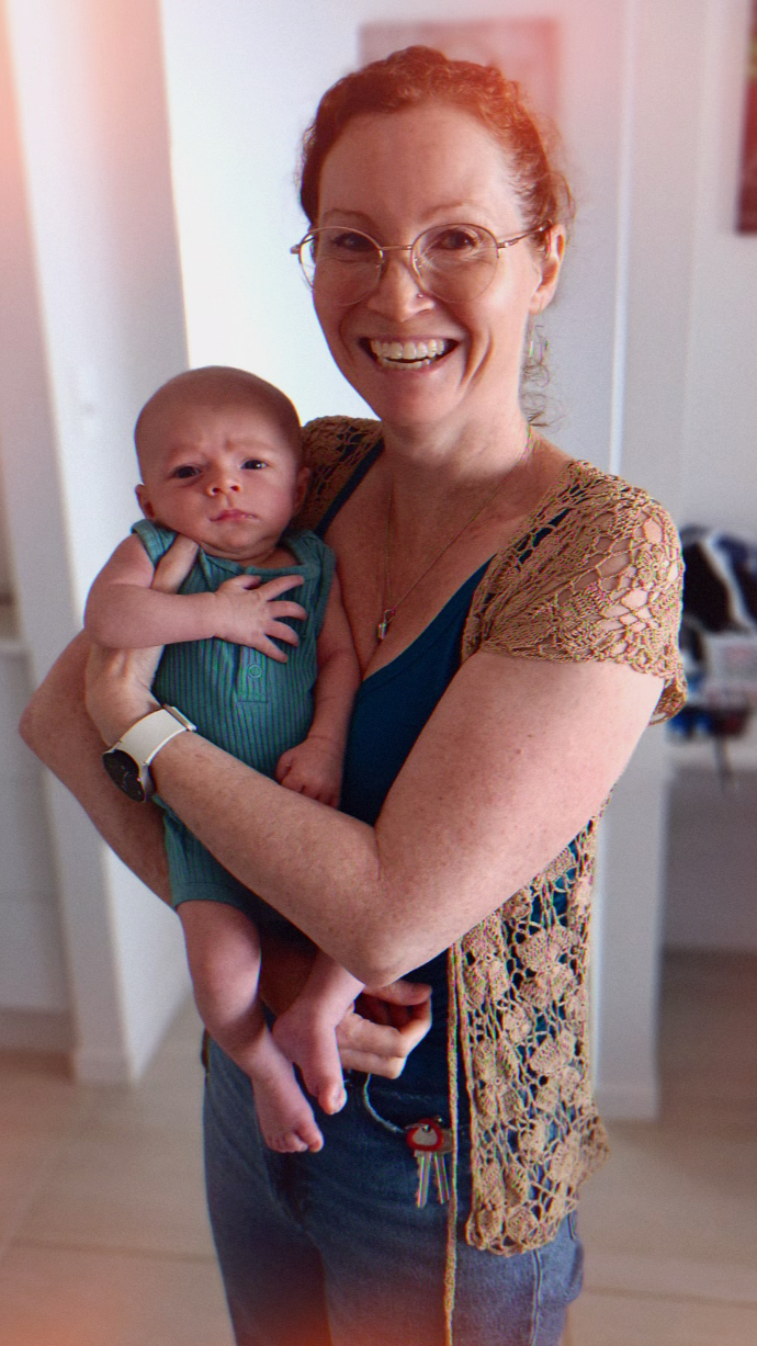 A smiling woman with red hair and glasses holding a baby with a serious expression, indoors in a home setting.