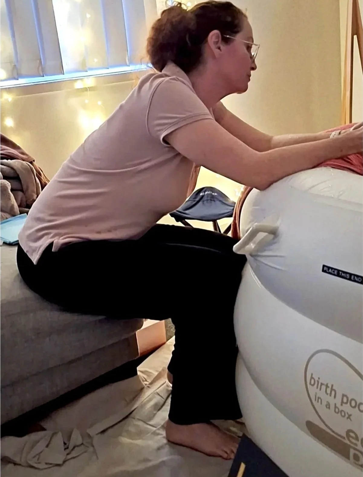 A woman with curly hair, glasses, and a pink shirt, sitting on a couch and leaning forward to examine a birth pod in a box.