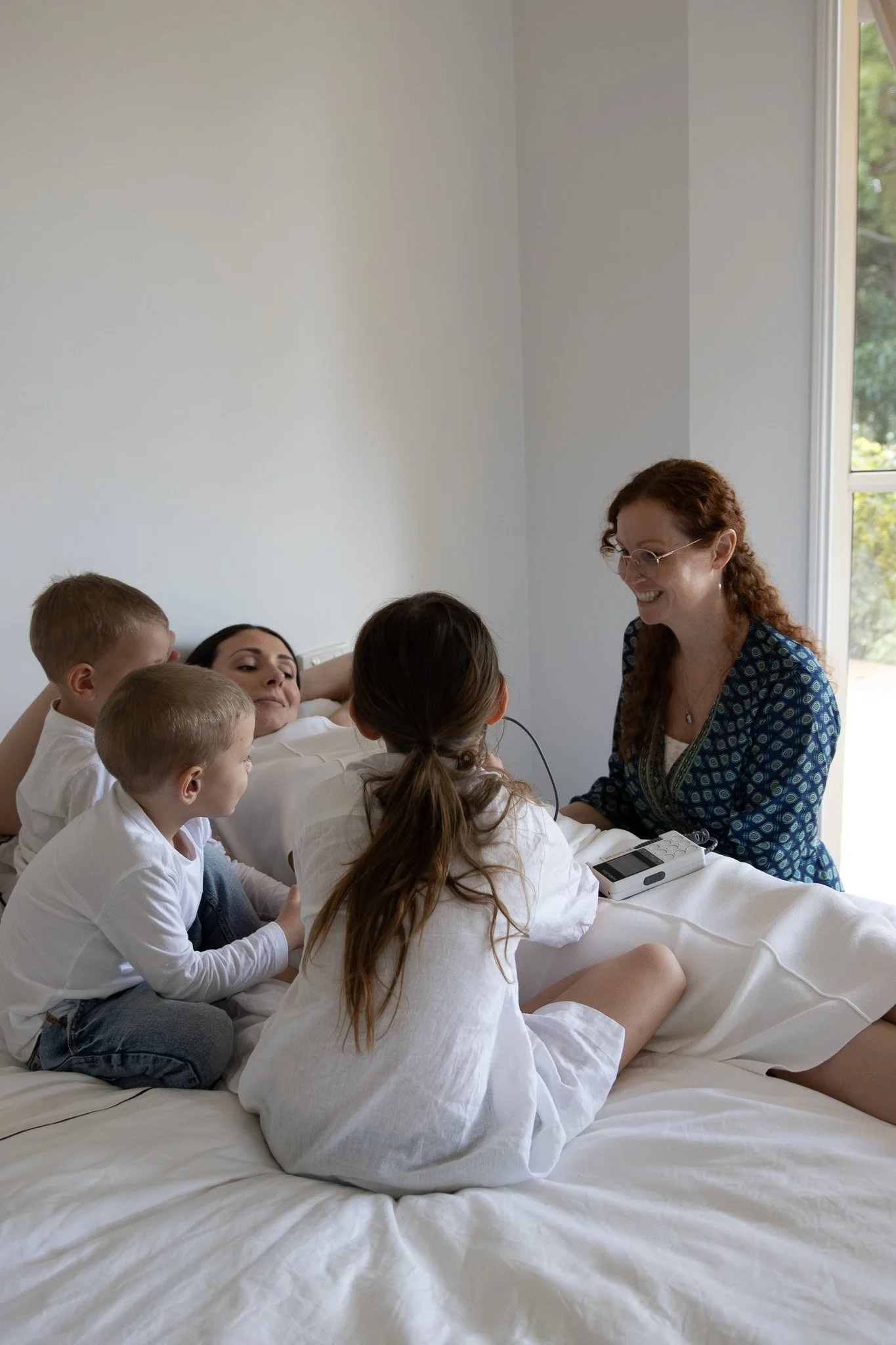 A woman lying on a bed with four children around her, smiling and talking to an older woman sitting nearby, in a bright room with a window.