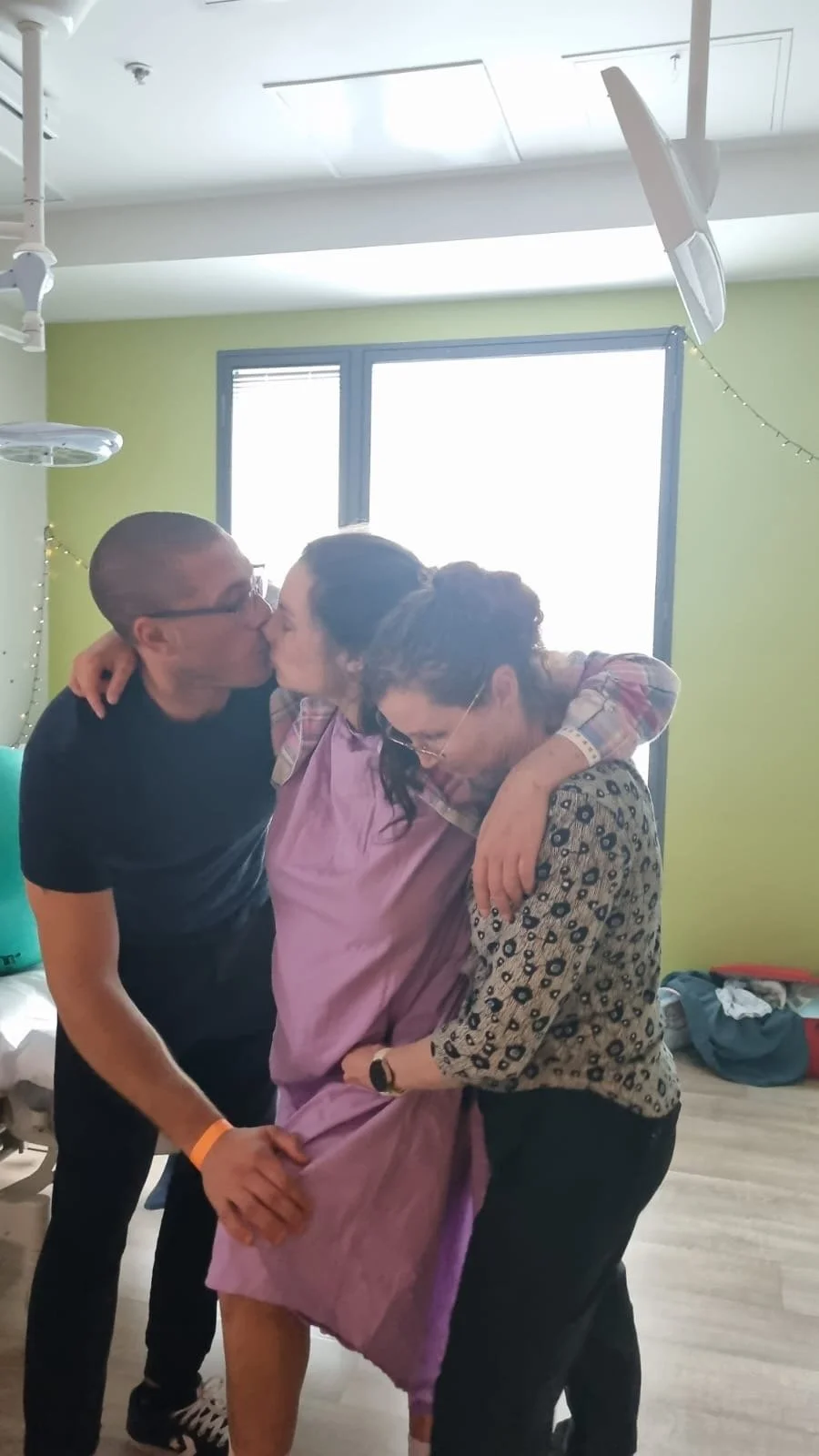 Two women and a man sharing a hug and kiss in a brightly lit room with green walls, a large window, and some personal items visible in the background.
