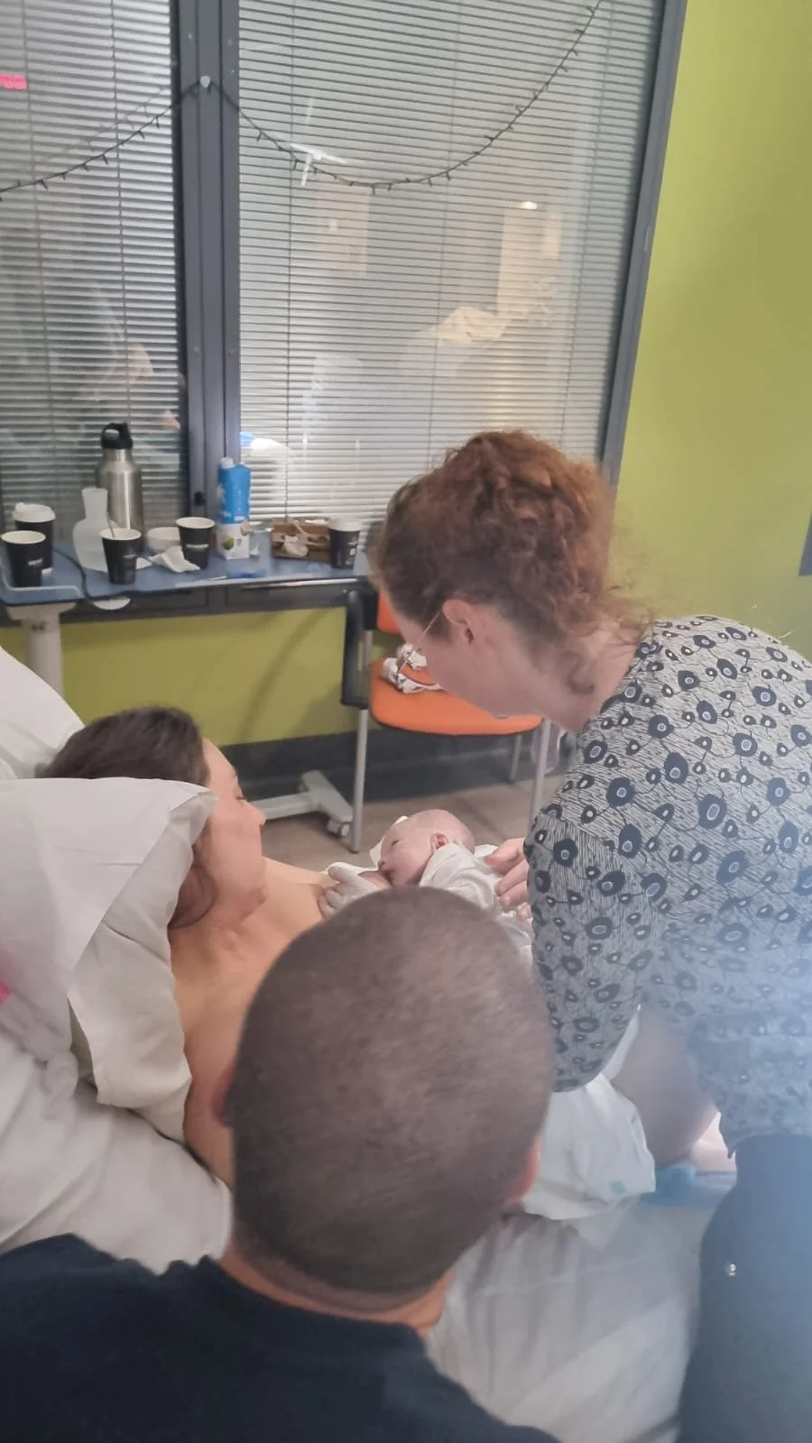 A woman in a hospital bed giving birth, with a healthcare worker holding her newborn baby, and a man sitting beside her in a hospital room with window blinds and cups on a table behind.