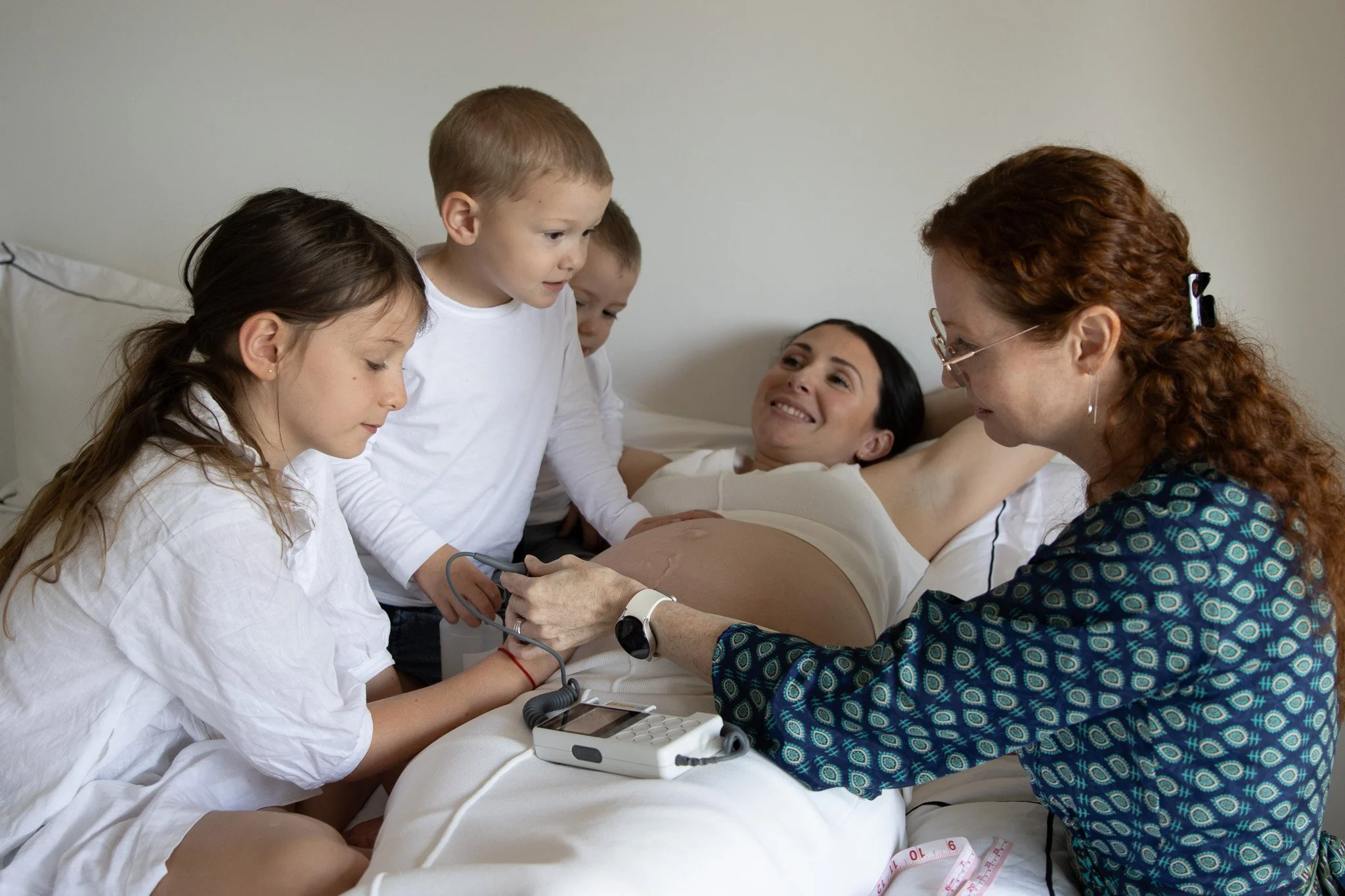 A pregnant woman lying in bed while a woman and three children check her heartbeat with a stethoscope and medical device.