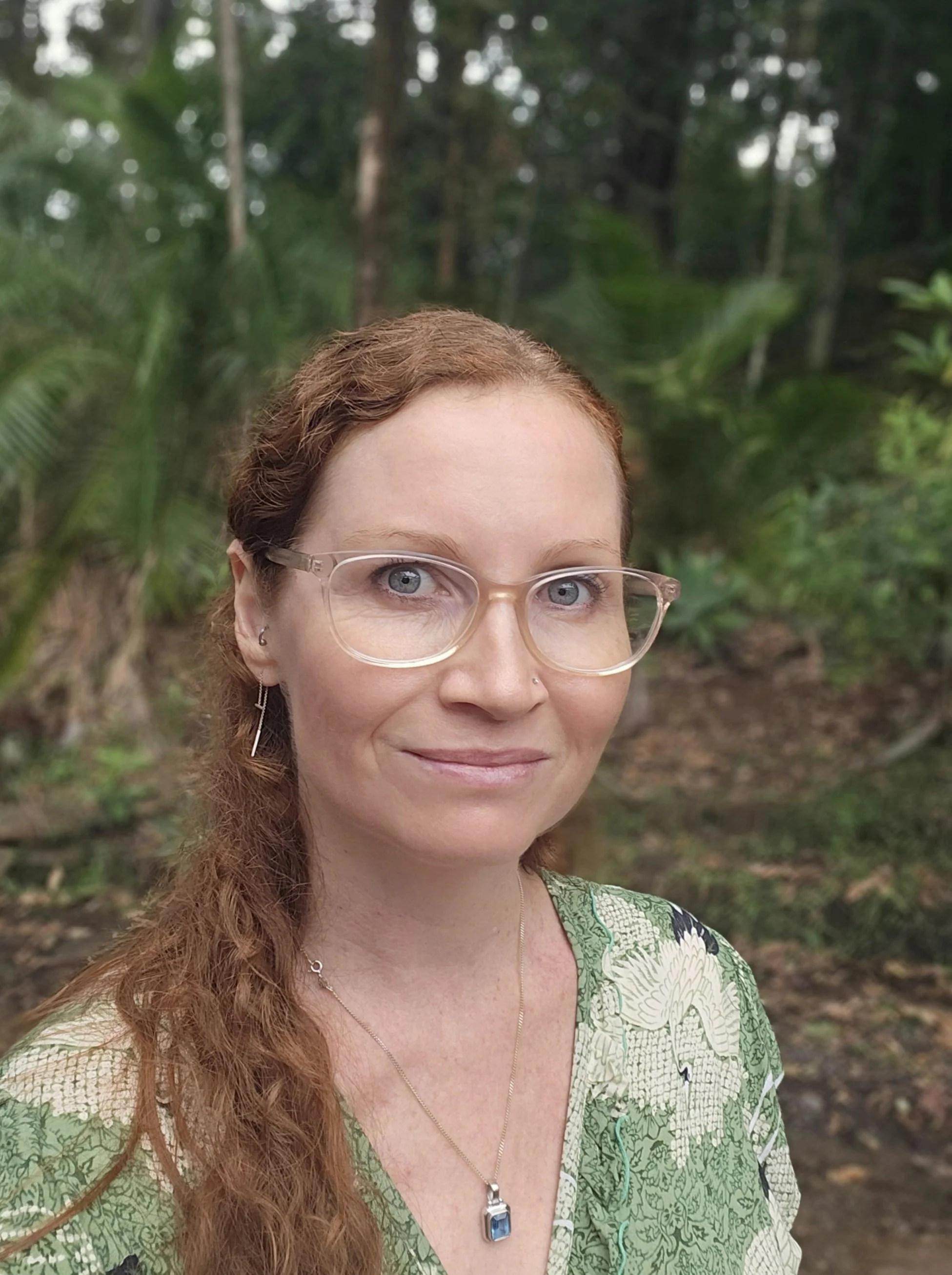 A woman with red hair, glasses, and a green patterned top standing outdoors in a forested area.