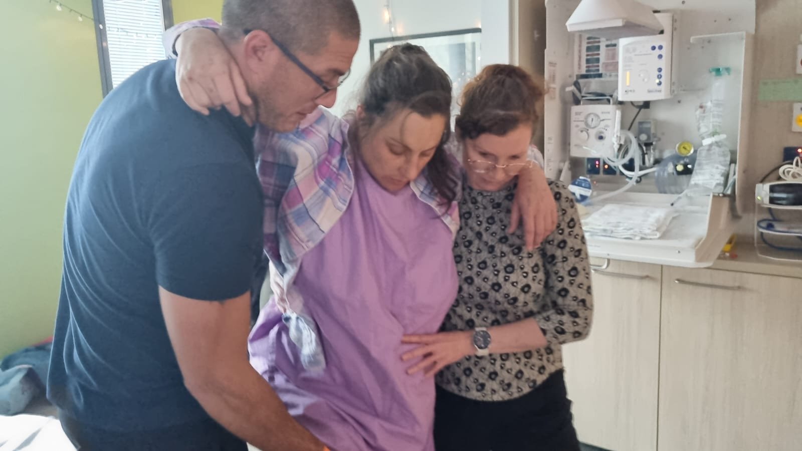 Three people supporting a woman in a hospital room, with medical equipment and supplies in the background.