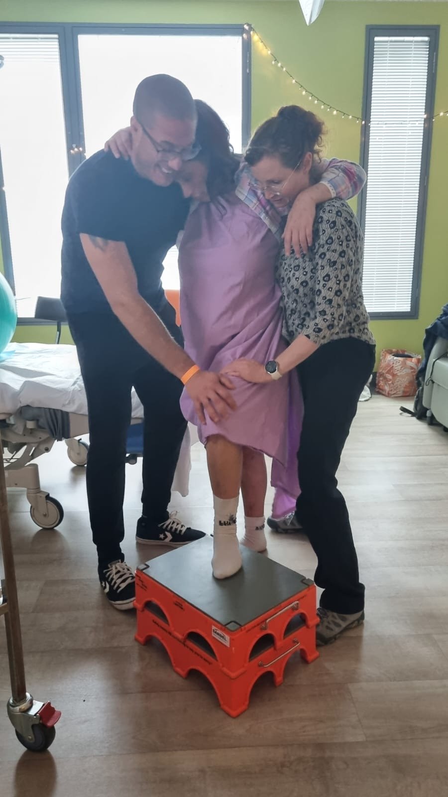A child in a hospital gown standing on a small orange platform, supported by two adults, during a celebratory moment with smiles and hugs in a brightly lit room.