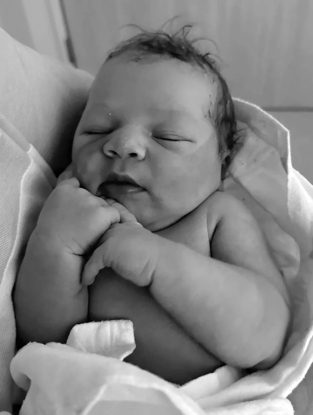A newborn baby sleeping peacefully, with fists near the face, wrapped in a cloth, in a hospital setting.