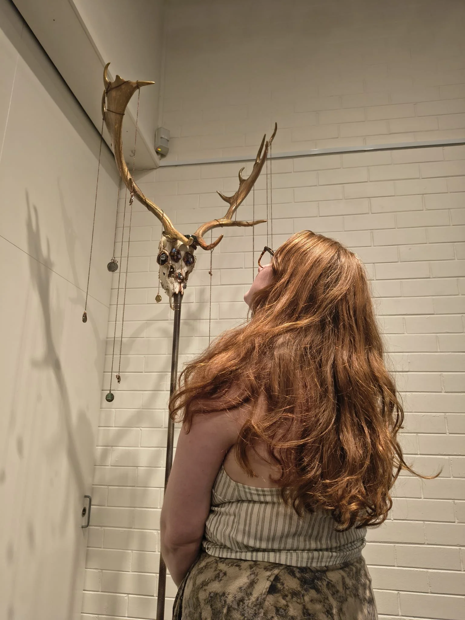 A woman with long curly red hair, wearing glasses, striped top, and patterned skirt, standing in a room with white brick walls. There's a mounted decorative deer skull with antlers decorated with jewelry hanging on the wall.