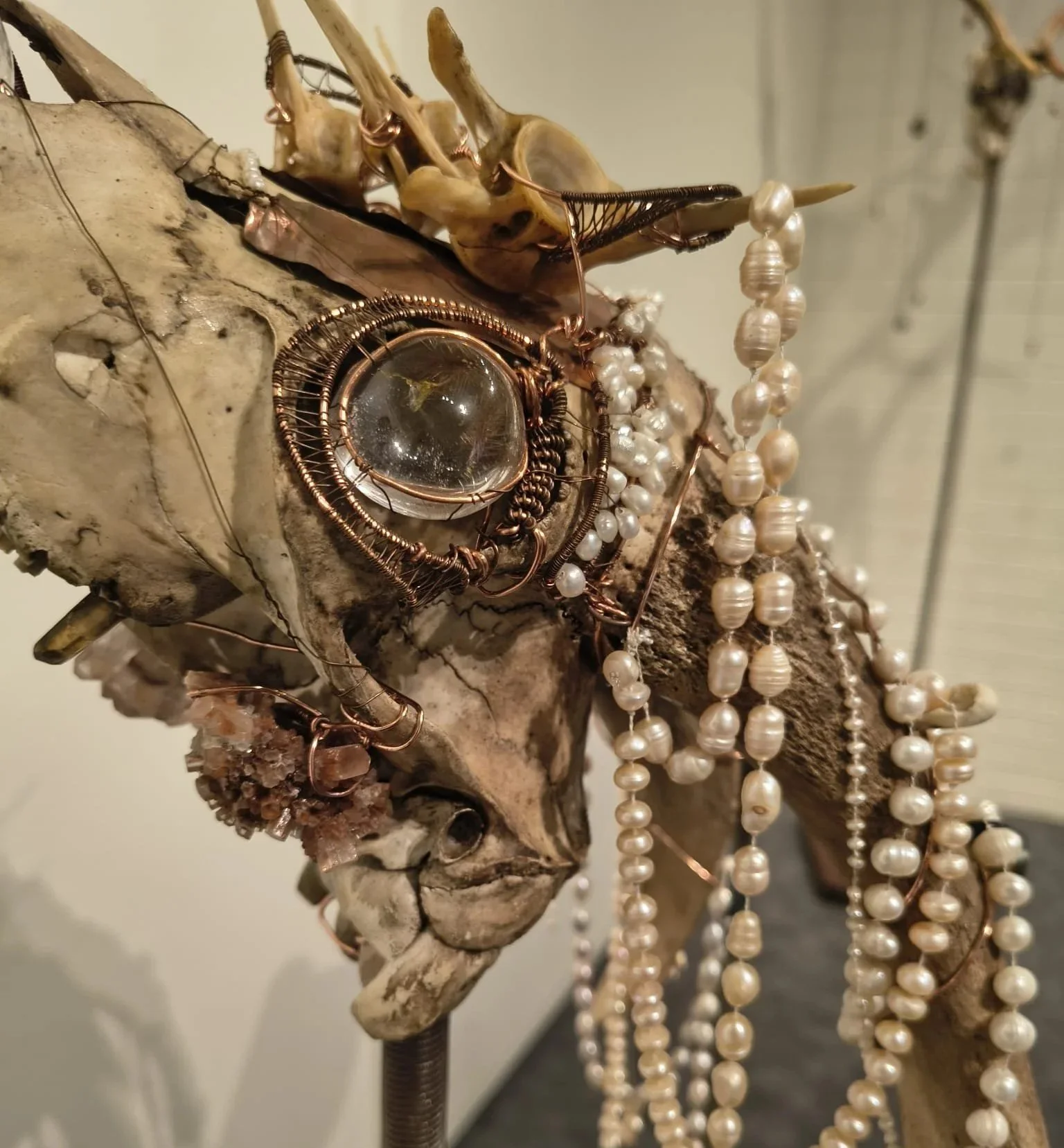 A display of jewelry including pearl necklaces, a copper wire-wrapped pendant, and various small rings, arranged on a piece of driftwood or weathered wood.