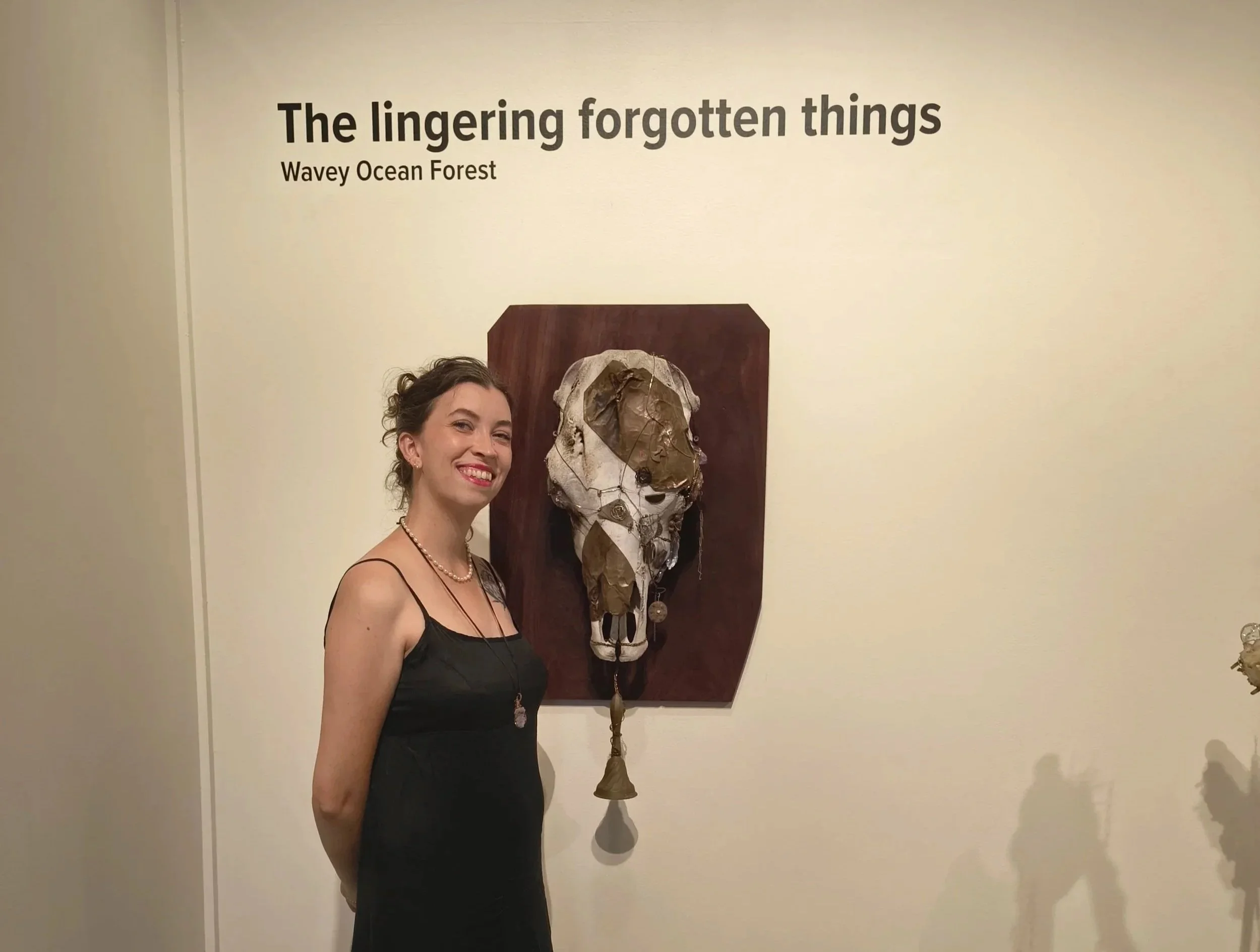 A woman with curly brown hair tied back, wearing a black dress and pearl necklace, standing in front of a wall with the text 'The lingering forgotten things, Wavey Ocean Forest,' and a display of a large, artistic skull with metallic and stone elements.