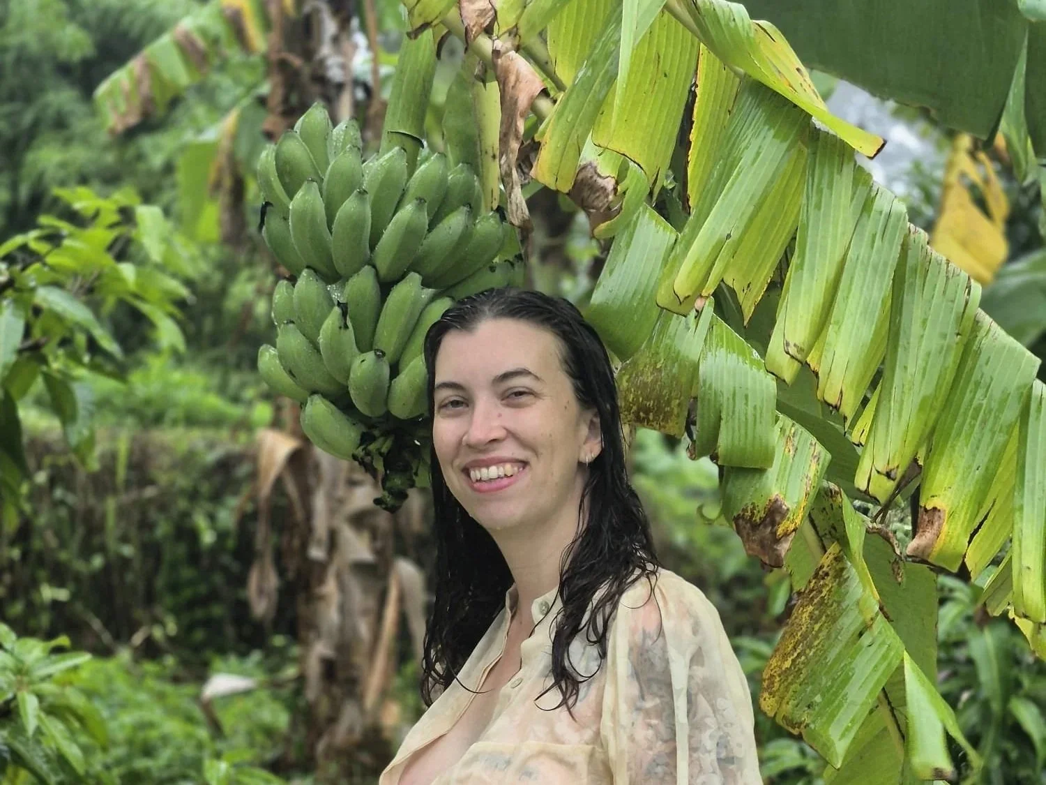 A woman with wet dark hair smiling in front of a large bunch of green bananas in a tropical setting.