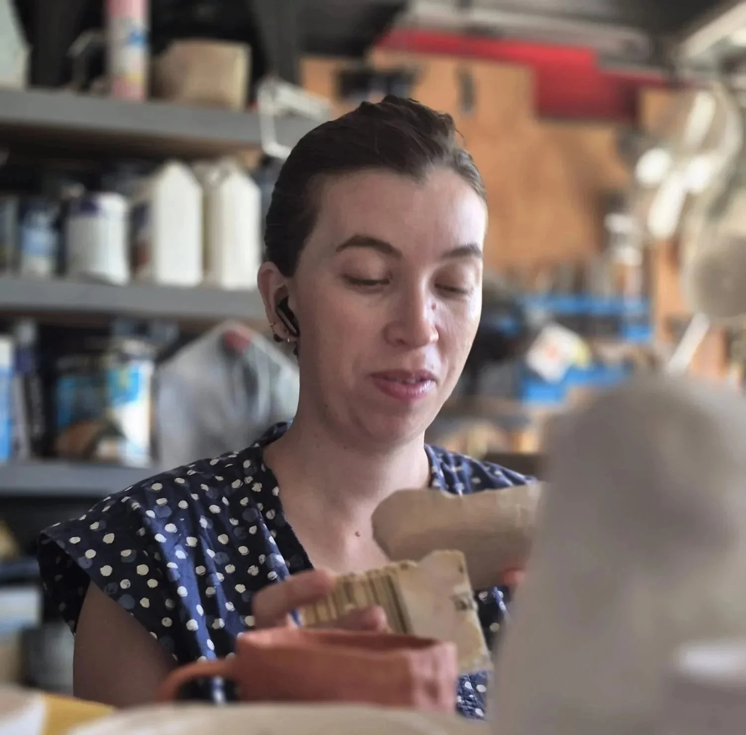 A woman with short dark hair, wearing a navy blue dress with white polka dots, looking at a piece of paper in a store or warehouse with shelves of supplies in the background.