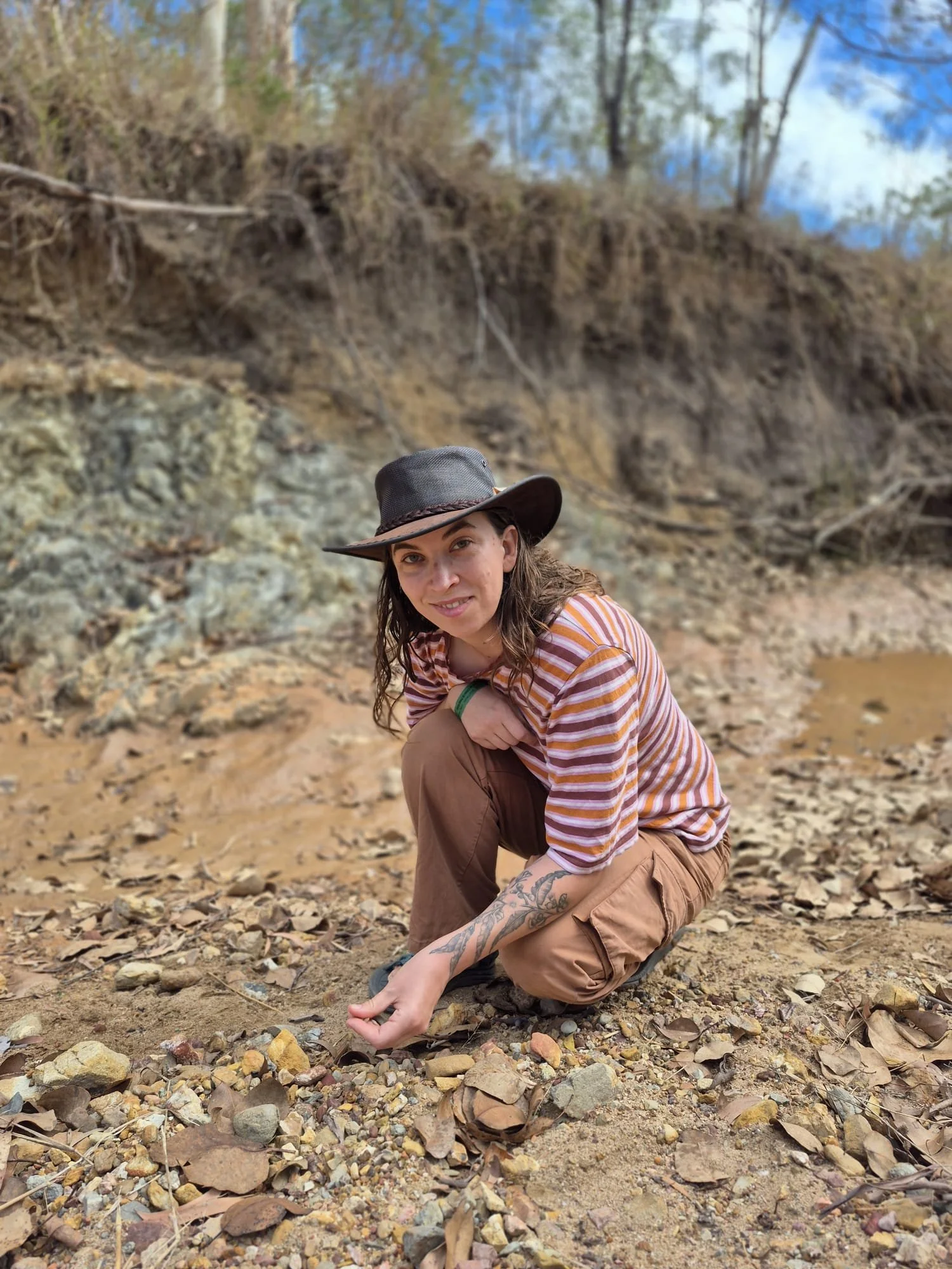 A woman in a striped shirt and tan pants crouches on rocky ground next to a river, wearing a wide-brimmed hat and smiling at the camera. The background shows a riverbank with trees and a partly cloudy sky.