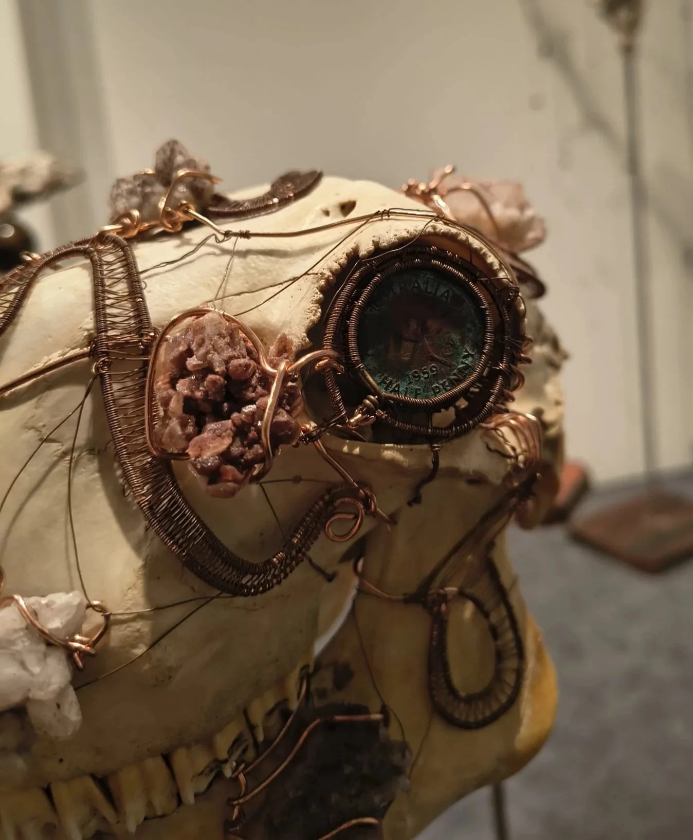 A human skull sculpture decorated with intricately wrapped copper wire, featuring a coin embedded in the area around the eye socket, and adorned with a small cluster of rocks or minerals near the nose.