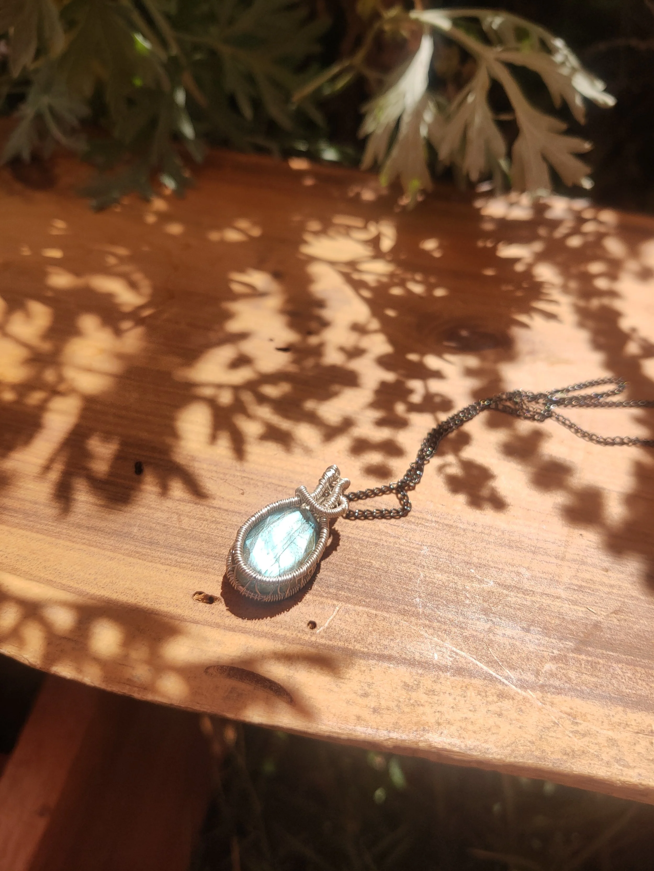 A silver necklace with an oval-shaped green gemstone pendant resting on a wooden surface. Shadows of leaves are cast on the surface and partially cover the necklace.