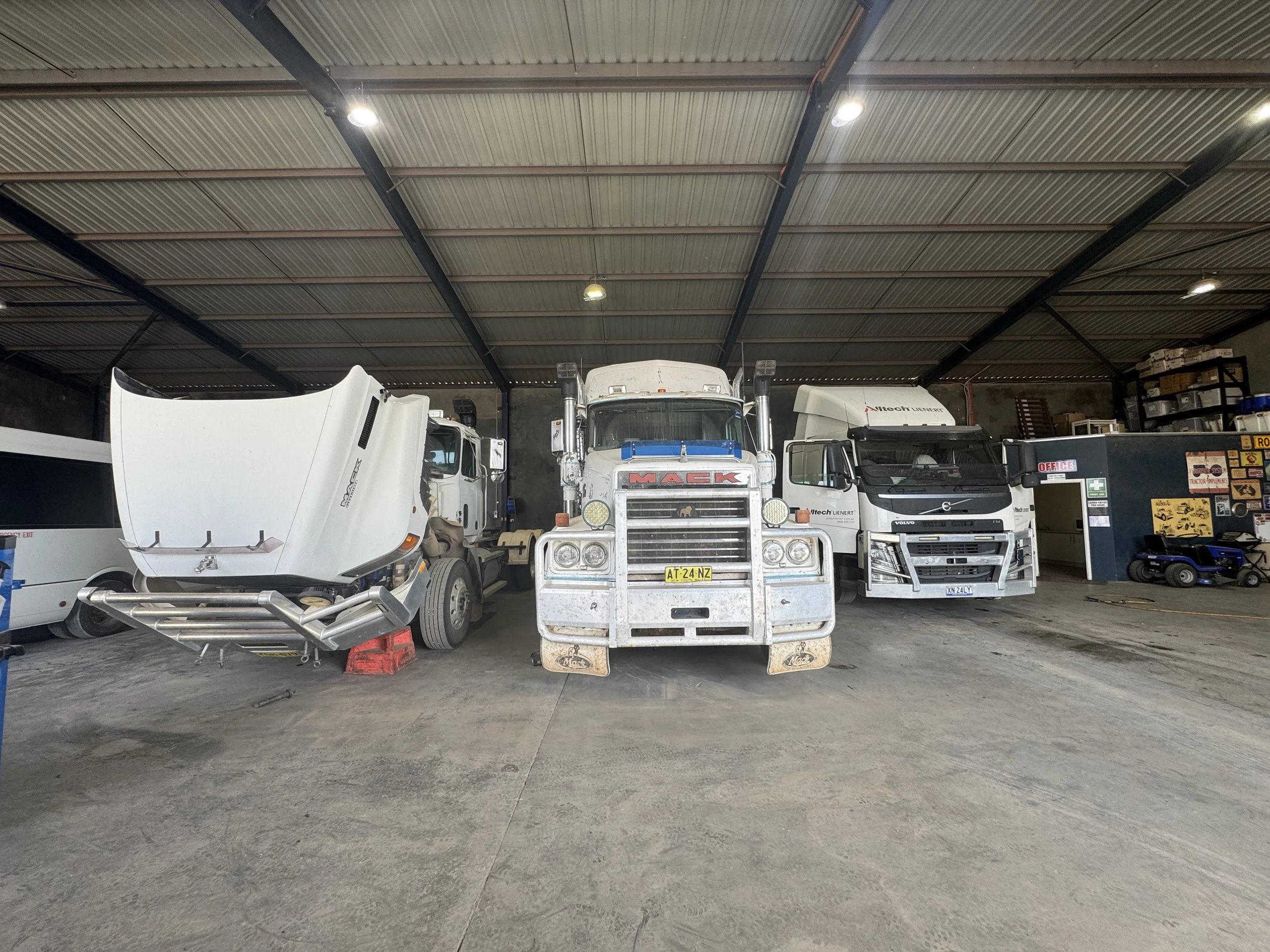 Garage with three trucks parked side by side, including a white truck with an open hood, a large white truck in the center, and a black truck on the right, inside a warehouse with shelving and tools.