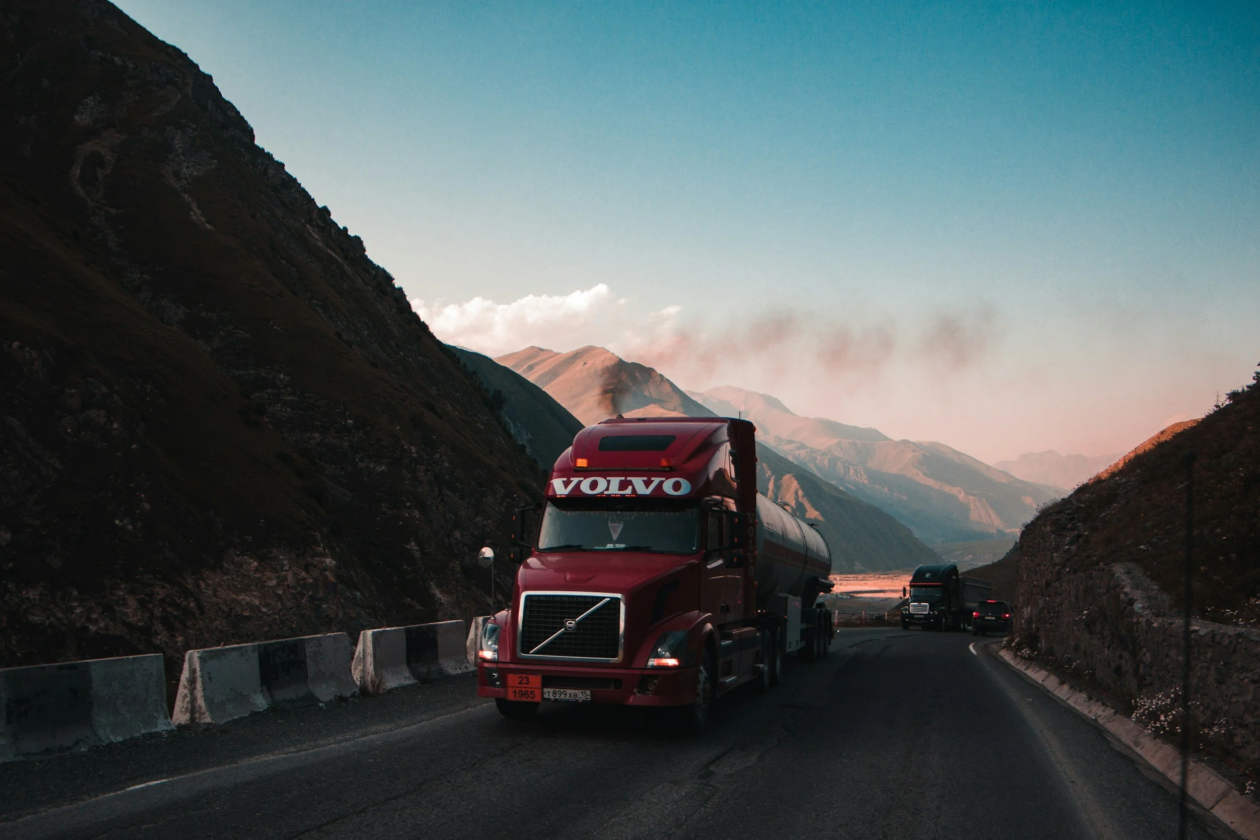 Red Volvo semi-truck driving on a mountain road at sunset with mountainous landscape and a few other vehicles in the background.