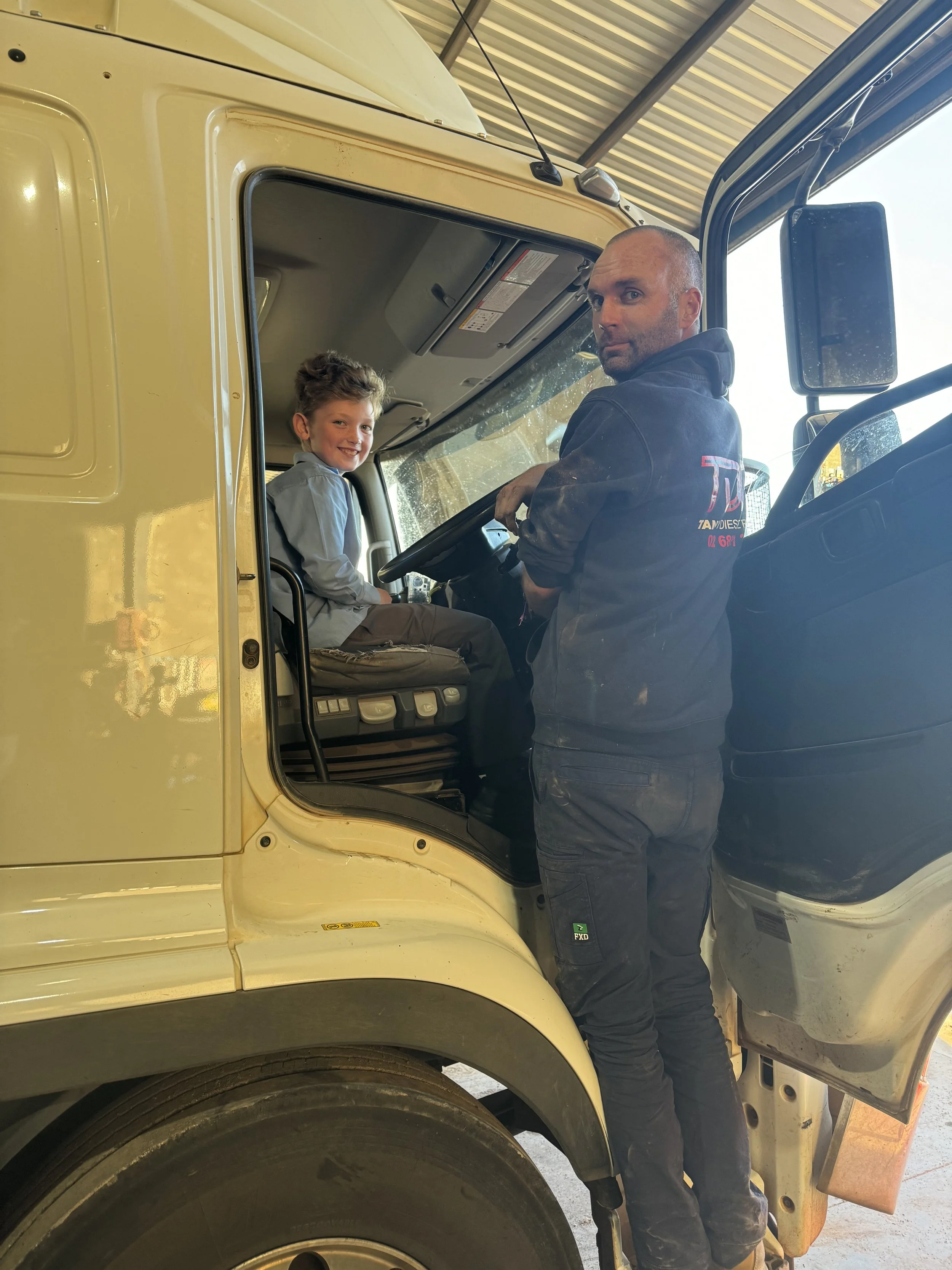 A smiling young boy sitting in the driver’s seat of a large yellow semi-truck, with a man standing beside him in the truck's cab, under a roofed structure.