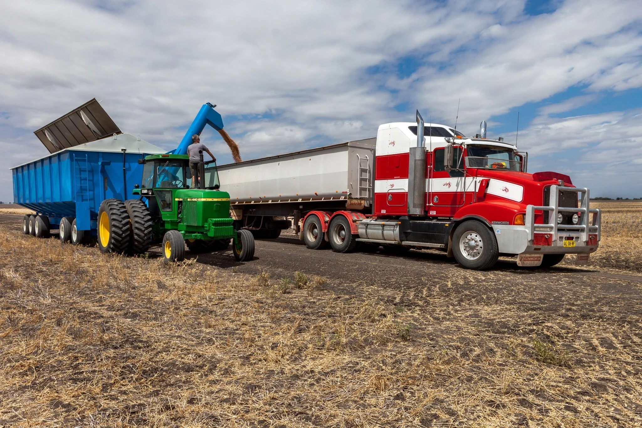 A farm scene with a large red and white semi-truck parked in a field, and a green tractor with large black tires unloading grain into a blue grain hopper attached to a trailer. The sky is cloudy with patches of blue.