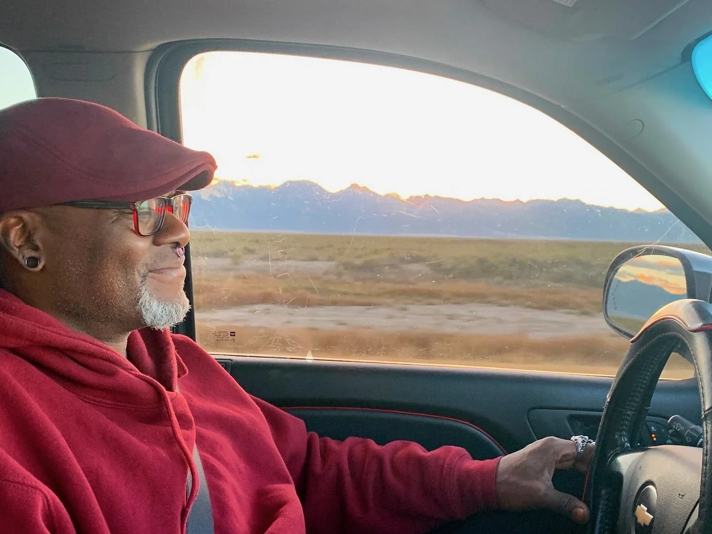 Man driving a vehicle through a desert landscape with mountains in the background during sunrise or sunset.
