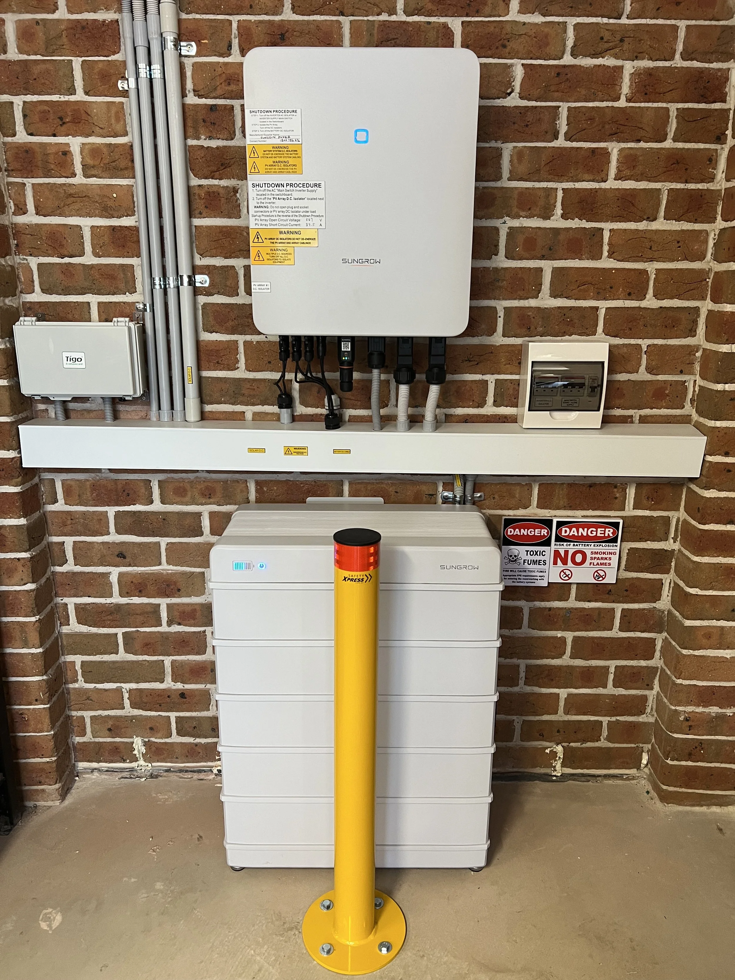 Sungrow solar power converter and battery system installed on a brick wall, with warning labels and a yellow safety bollard.