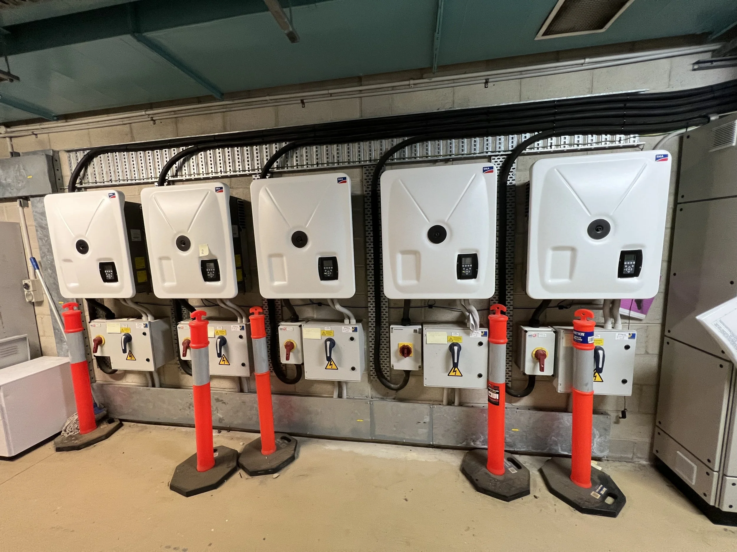 Row of wall-mounted electrical inverters with warning signs and orange safety barriers in front.