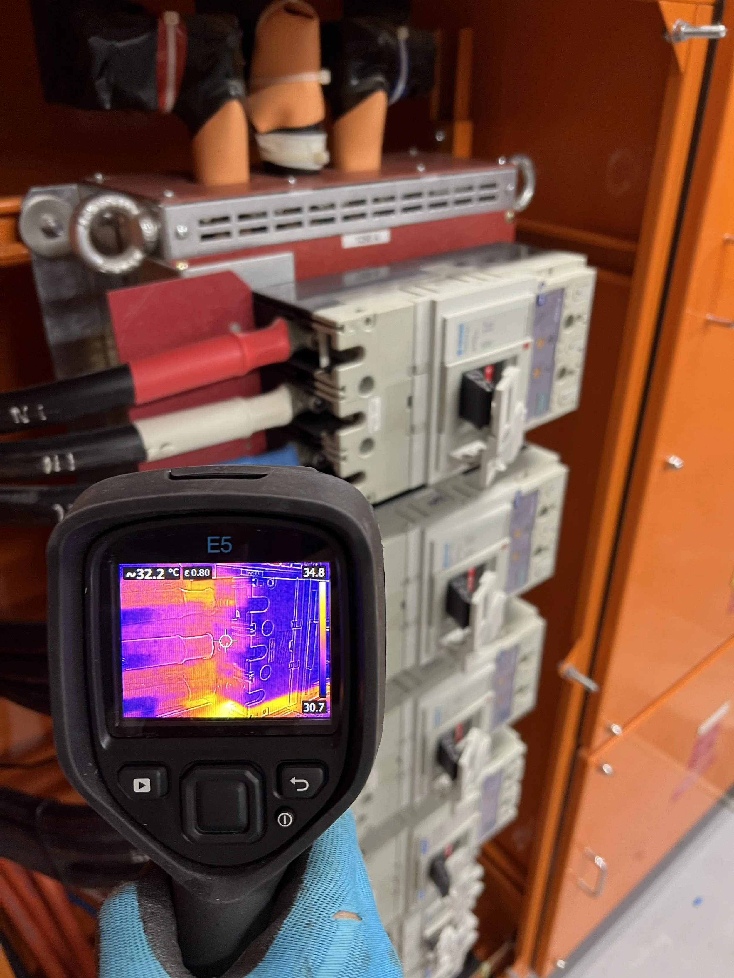 Person holding a thermal imaging camera in front of electrical switchgear, displaying temperature readings on the screen in a panel room.