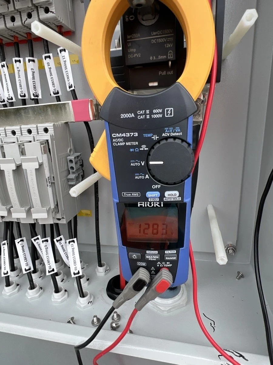 Digital clamp meter measuring electrical current in a control panel, with labeled wires and circuit components visible.