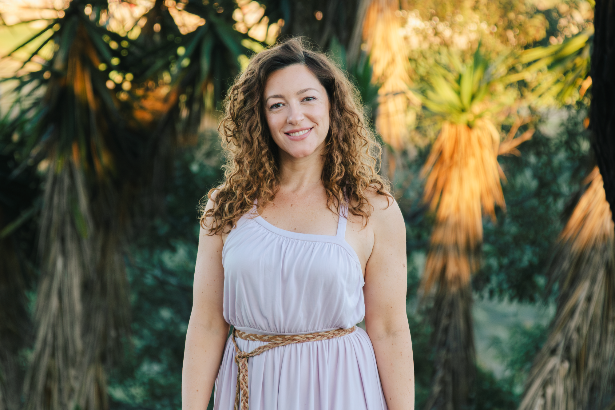 A woman with curly brown hair smiling outdoors, wearing a white dress with a beige woven belt, with tropical plants in the background.