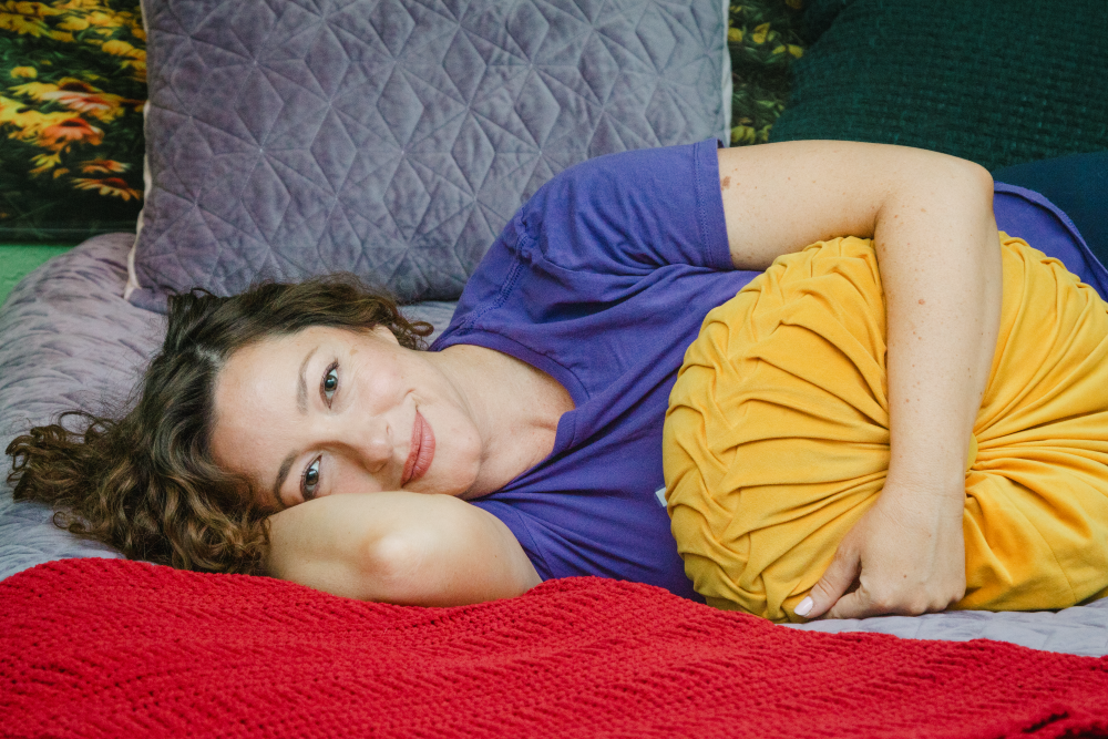 Woman with curly hair laying on her side, resting her head on her arm, surrounded by colorful pillows and blankets.