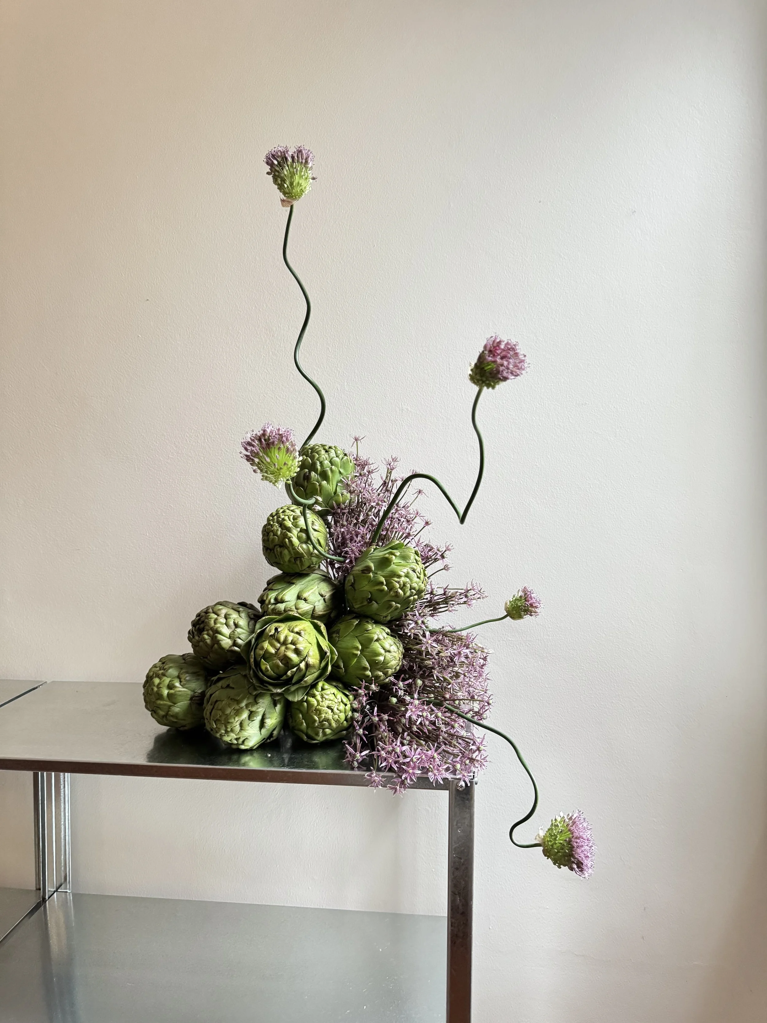 A floral arrangement with artichokes, purple flowering plants, and pink allium flowers on a metal table against a plain beige wall.