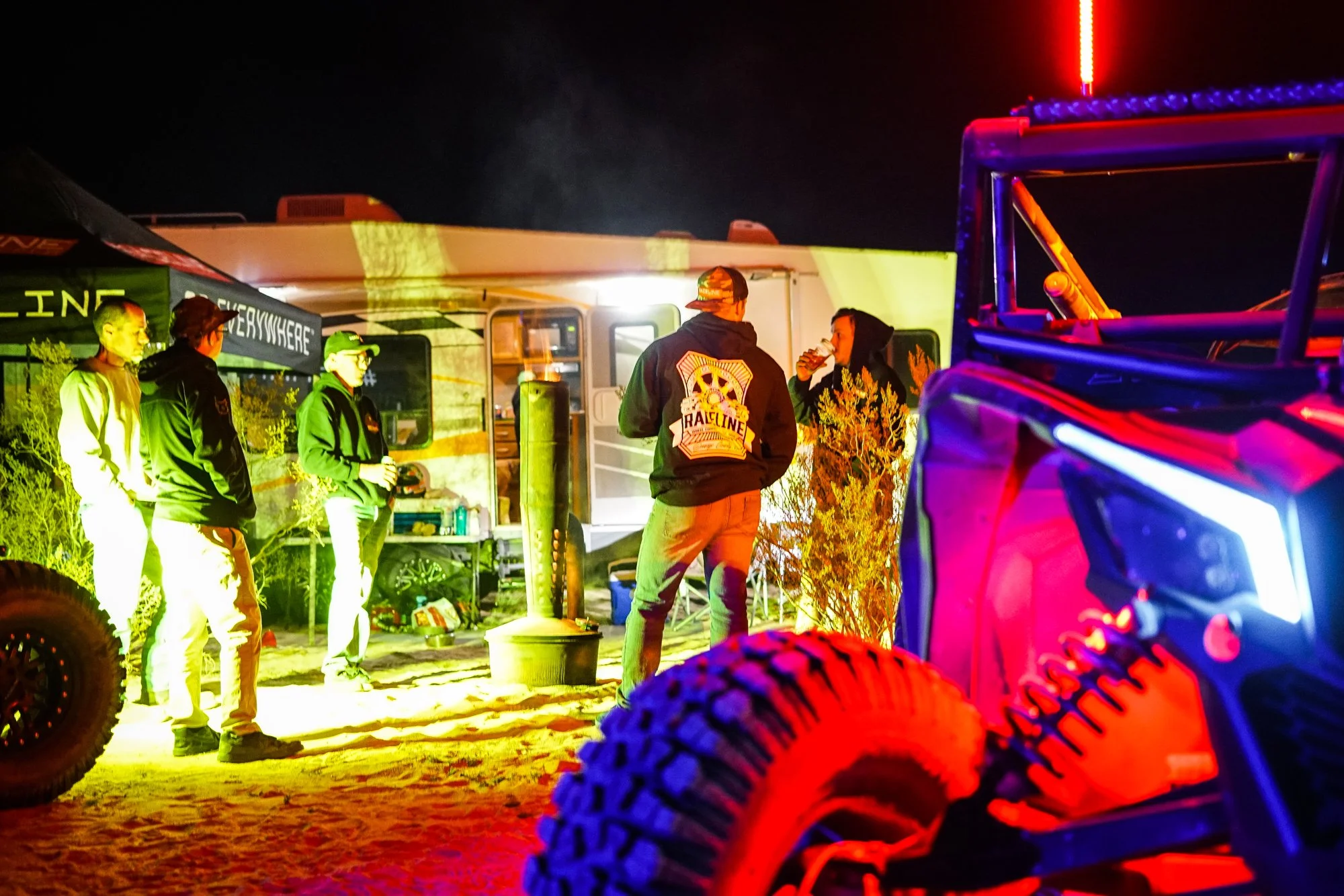 Group of people standing outdoors at night, illuminated by colorful lights, near RV with a flag, off-road vehicle with large tires, and campfire setup.