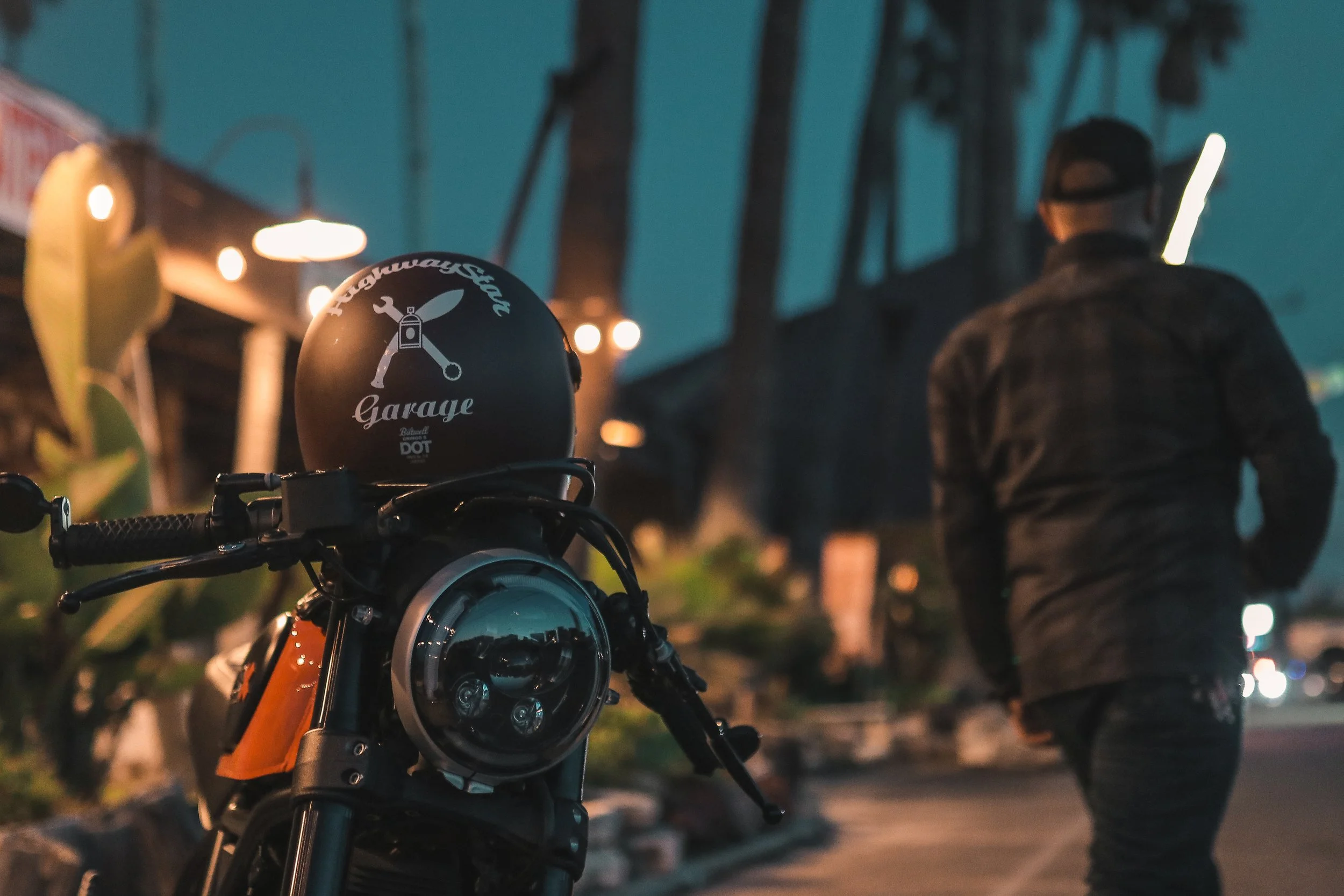 A motorcycle with a black helmet on the seat, parked outside a garage at dusk, with a person wearing a black jacket and cap walking away in the background.