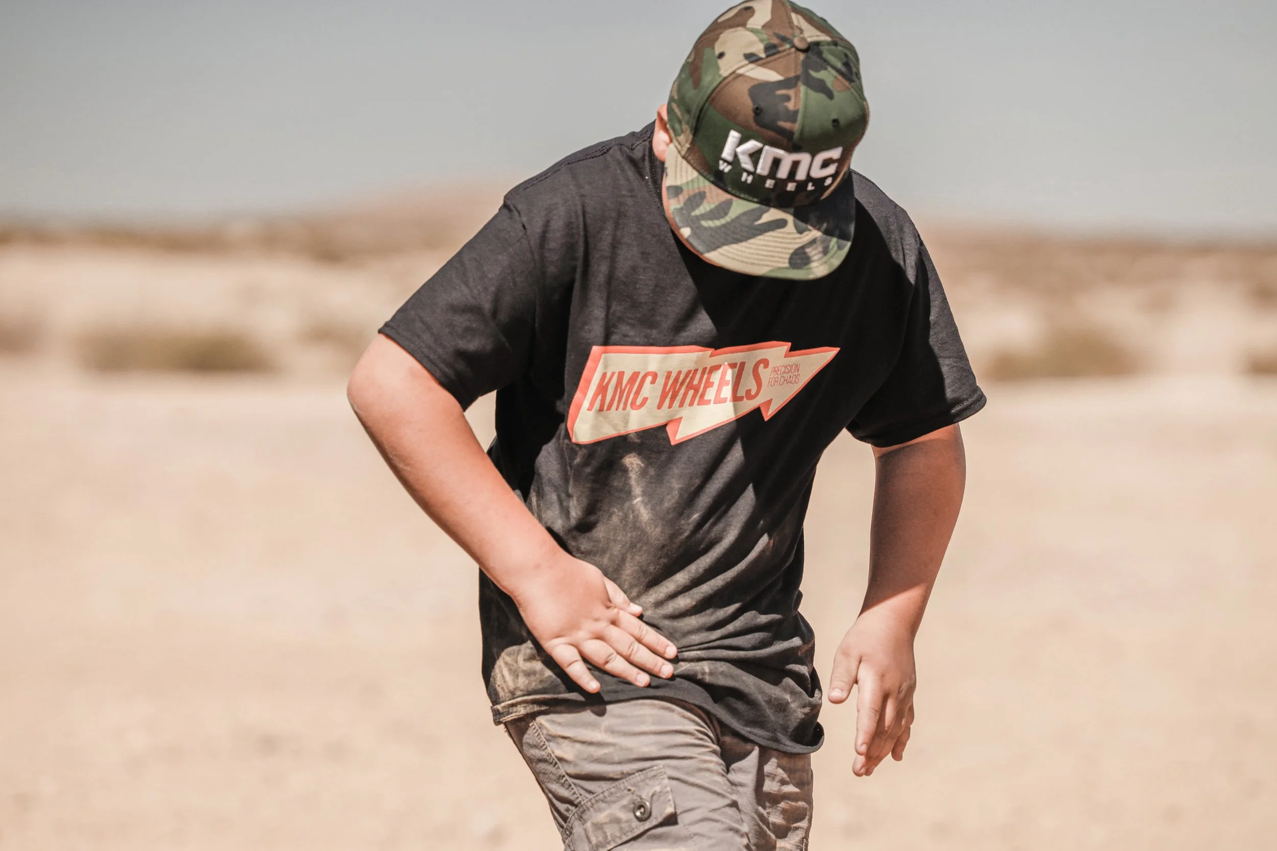 A person wearing a camouflage cap and a black T-shirt with a red and white logo, standing on a sandy terrain with a blurred background.