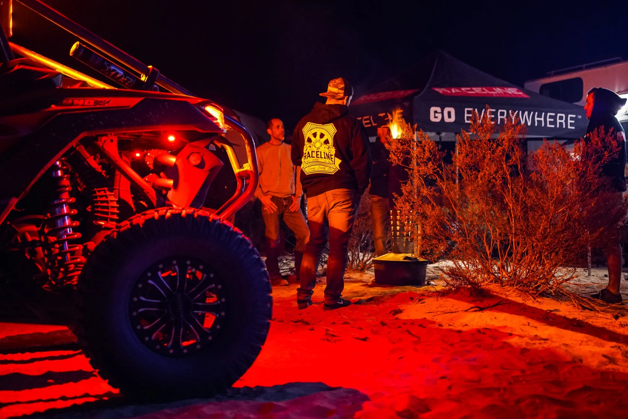 A nighttime scene with a remote-controlled off-road vehicle and several people gathered under a tent with the text 'Go Everywhere' and 'Raceline' on it. The scene is illuminated by red light, and the background appears to be a sandy or desert area.