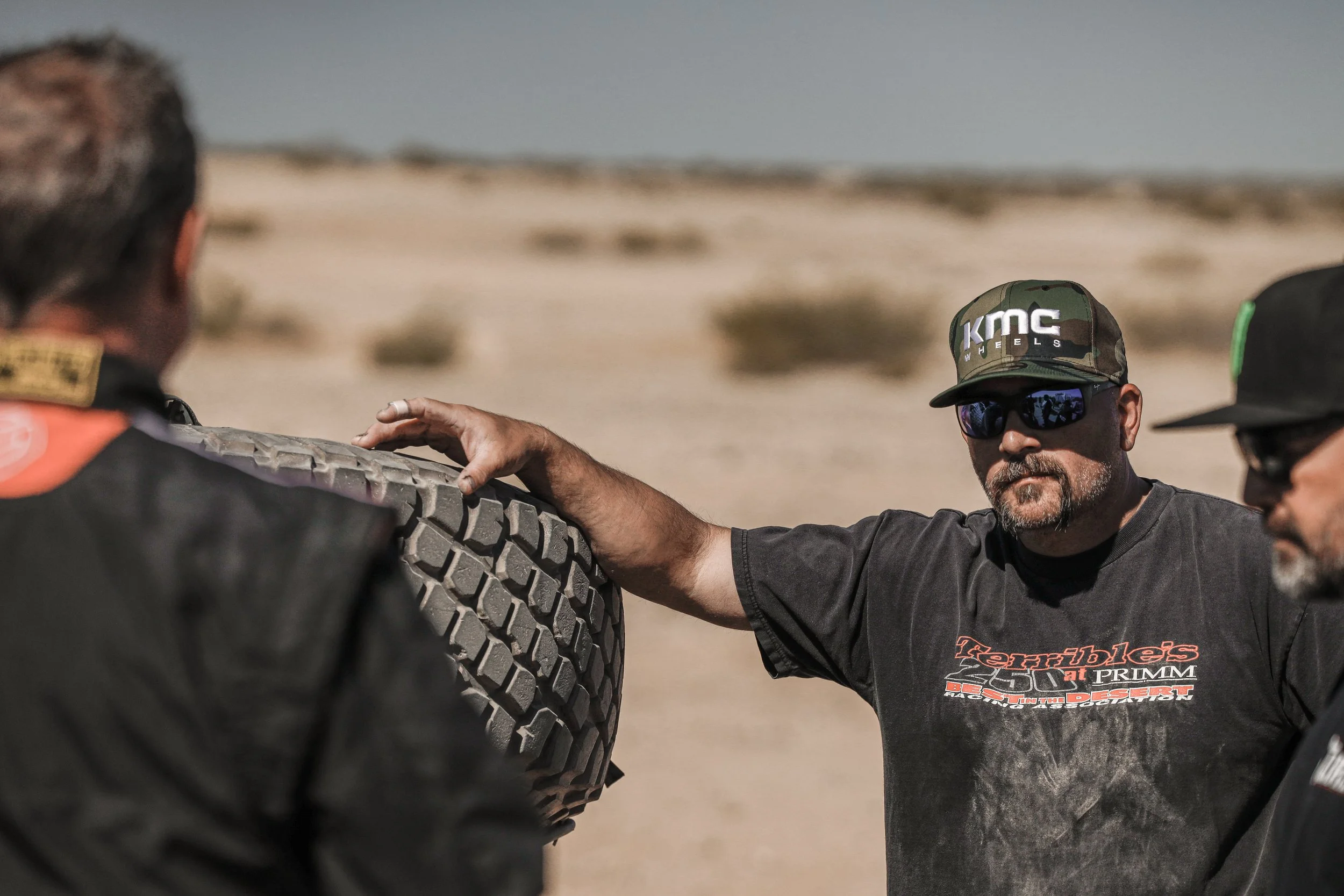 Three men in a desert, one wearing a camouflage cap and sunglasses, inspecting a tire.