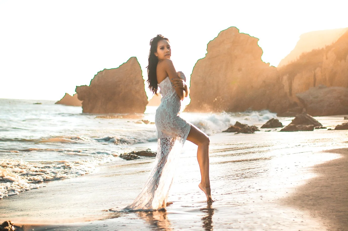 A woman in a lace dress standing at the shoreline with ocean waves and large rocks and cliffs in the background during sunset.