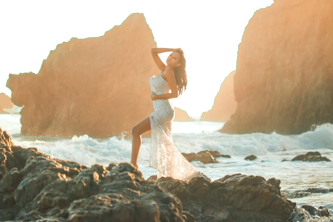 A woman in a white lace dress stands on rocks at the beach during sunset, with large rock formations in the background.