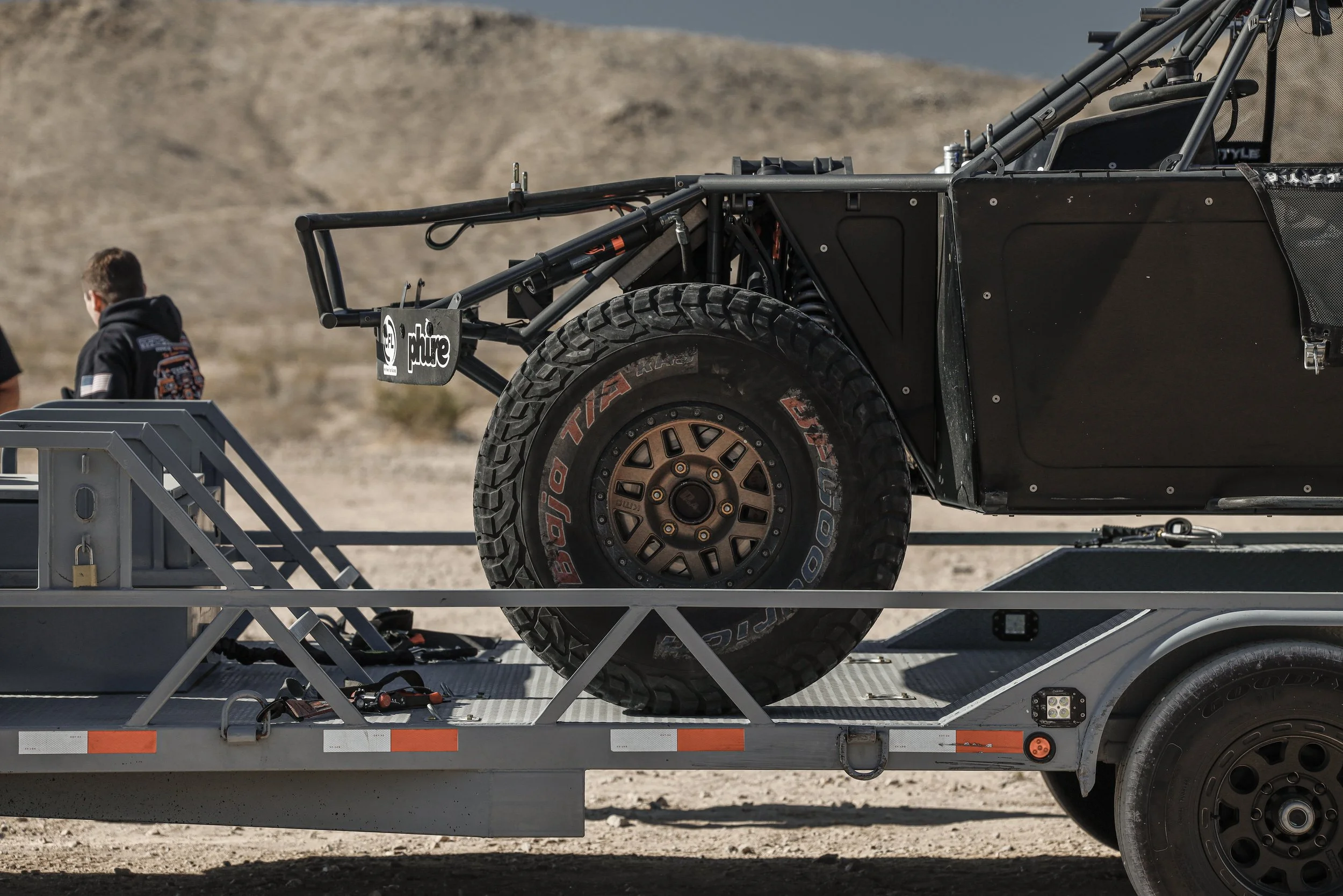 A black off-road racing vehicle on a flatbed trailer in a desert landscape, with racing tires and a roll cage visible.
